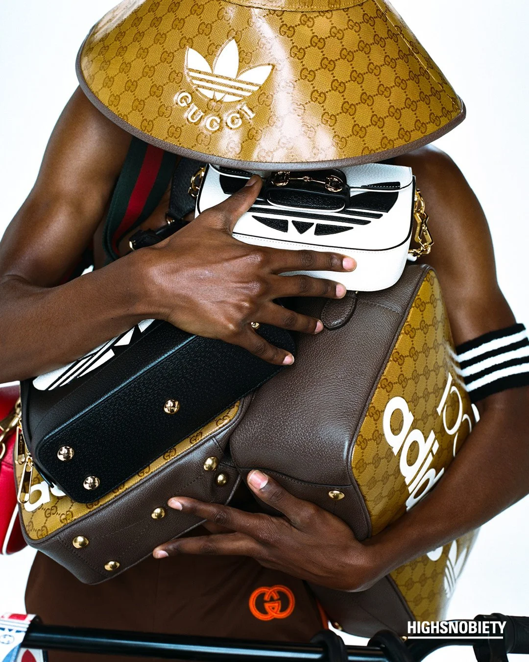 Person holding several designer handbags and wearing a large gold Gucci hat with the Adidas logo. The person's face is not visible, and they are dressed in casual attire.
