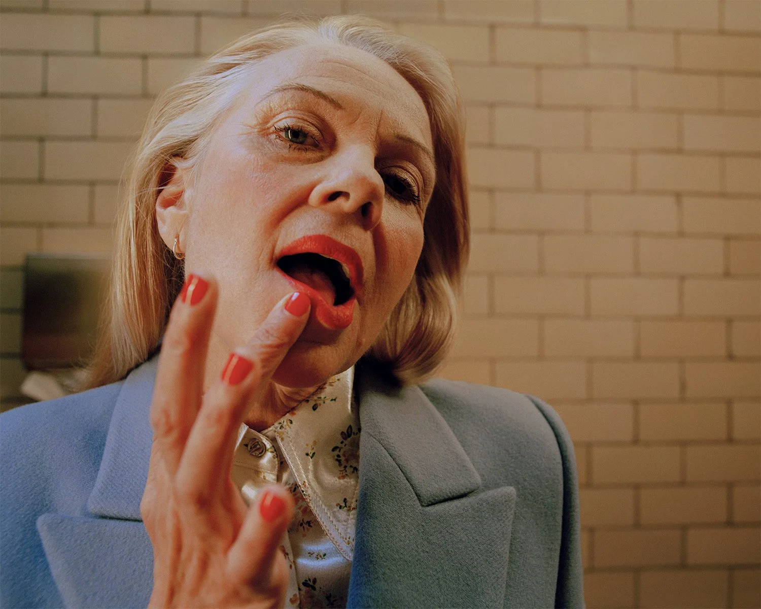 Close-up of an older woman with blonde hair, red lipstick, and orange nail polish, wearing a light blue blazer and floral blouse, touching her lips with her fingers against a brick wall background.