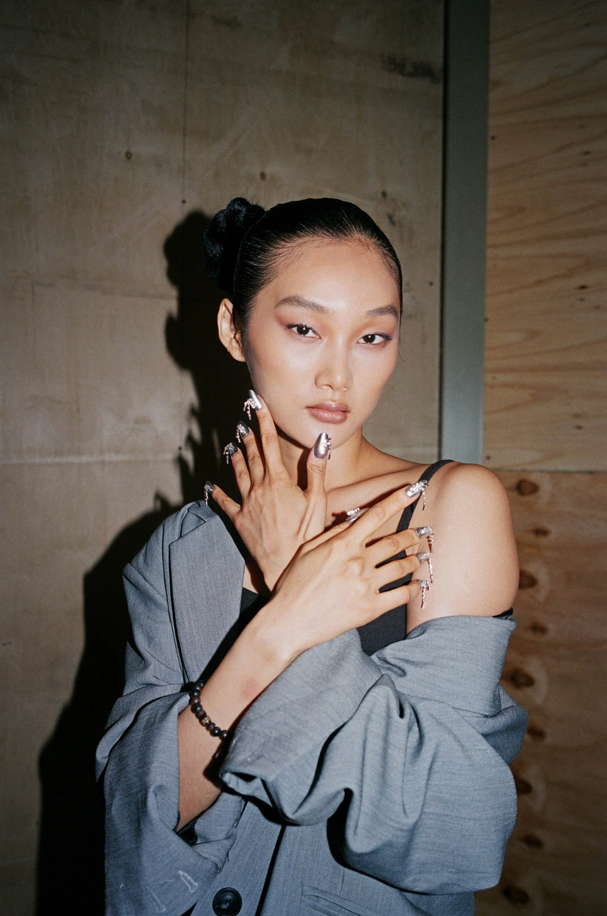 A young woman with black hair styled in a bun, wearing a gray blazer and black top with thin straps, posing with her hands near her face. She has metallic manicure nails and jewelry, with one shoulder slightly exposed.
