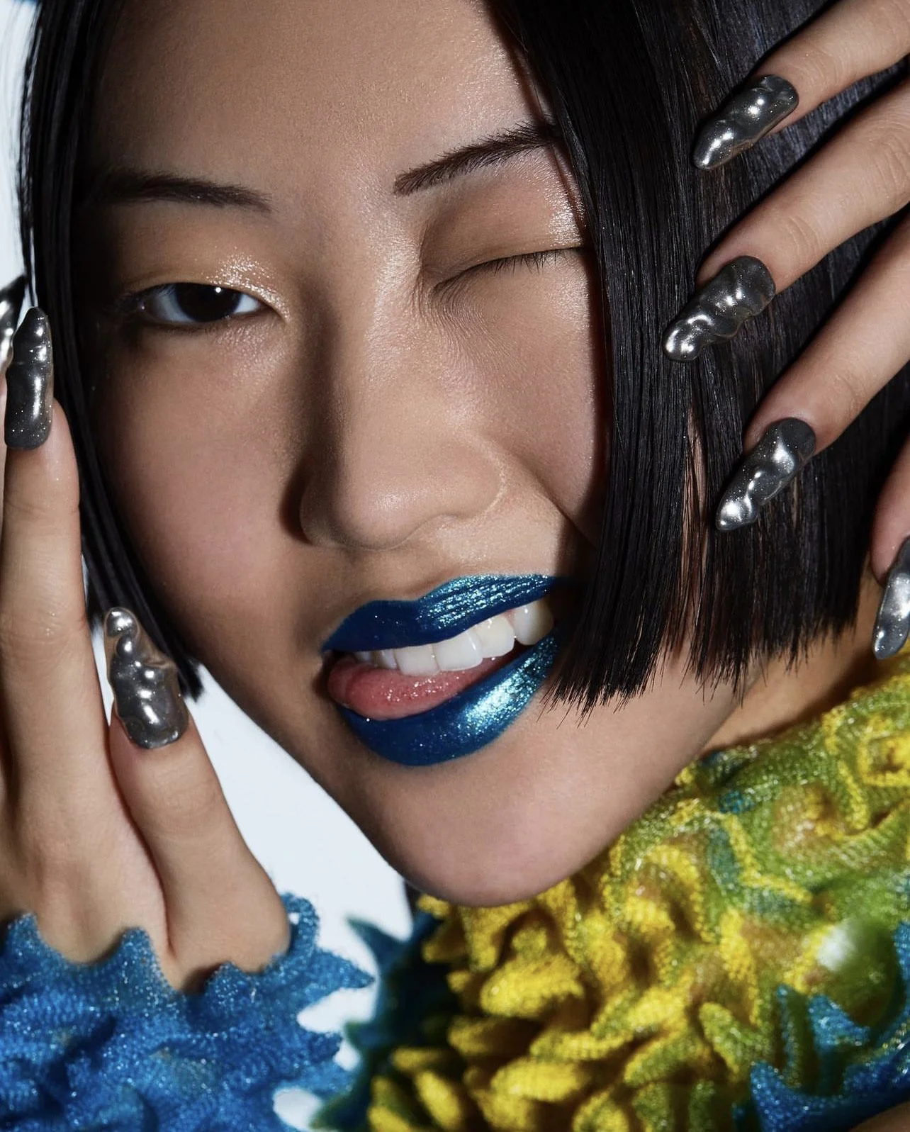 Close-up of a woman with short black hair, one eye closed, sticking out her tongue, wearing metallic blue lipstick, and holding her face with her fingers. She has long, metallic, silver nails and a colorful, textured garment.
