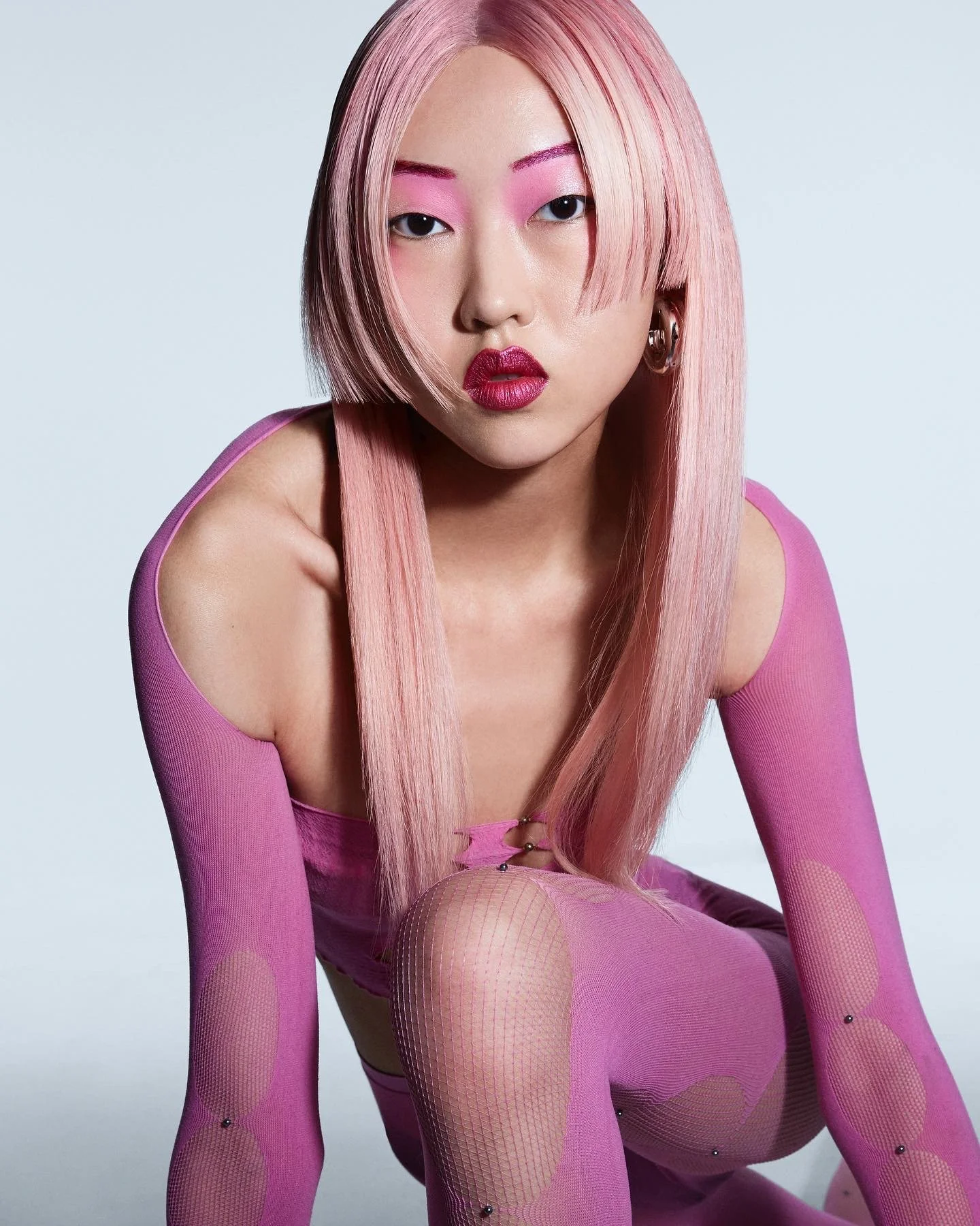 A woman with long pink hair and pink makeup, wearing a pink cut-out bodysuit and fishnet tights, sitting in a crouching pose against a plain background.