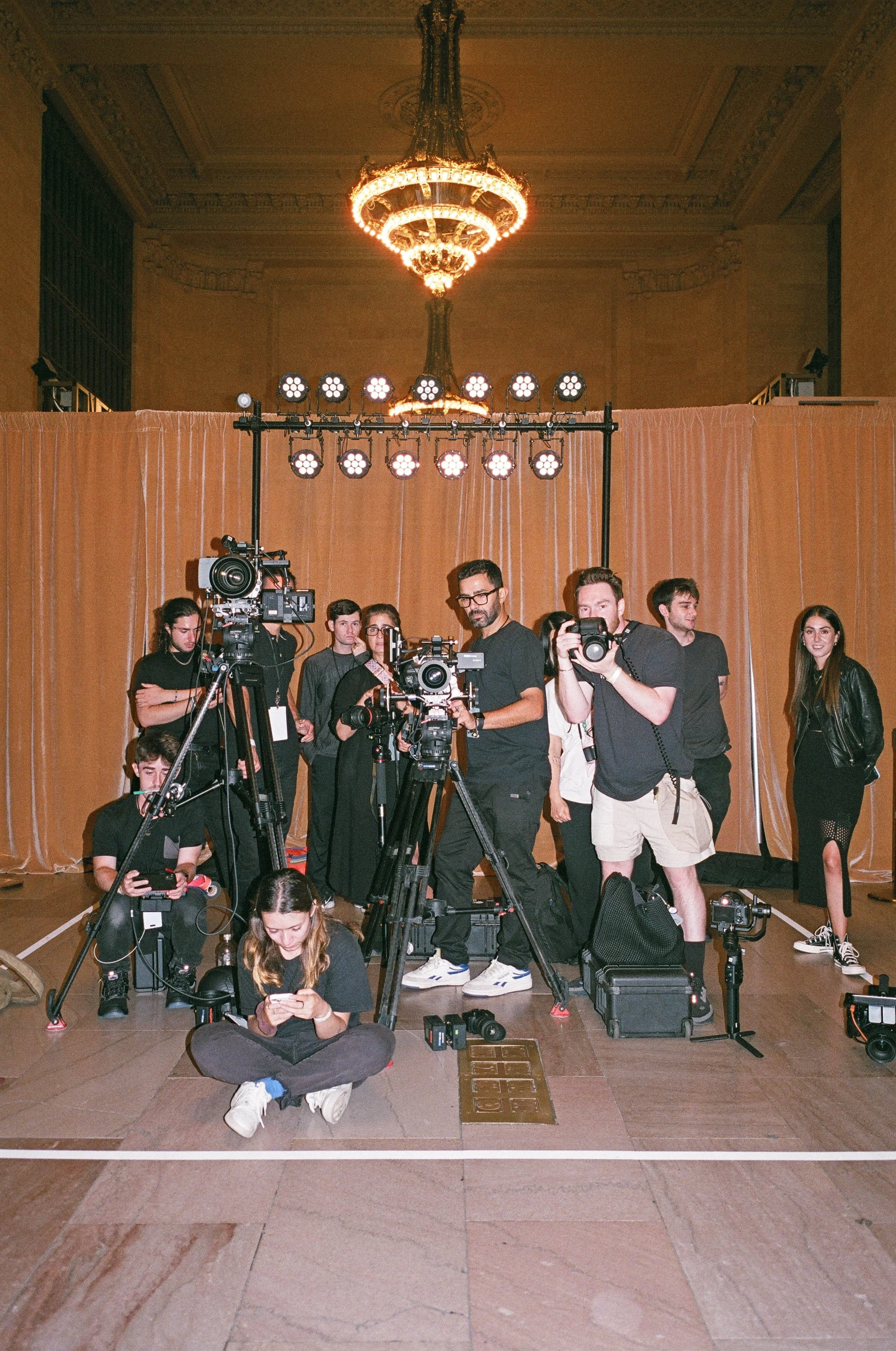 Group of photographers and videographers preparing for a shoot in a grand room with a chandelier, cameras set on tripods, and a brown curtain backdrop.