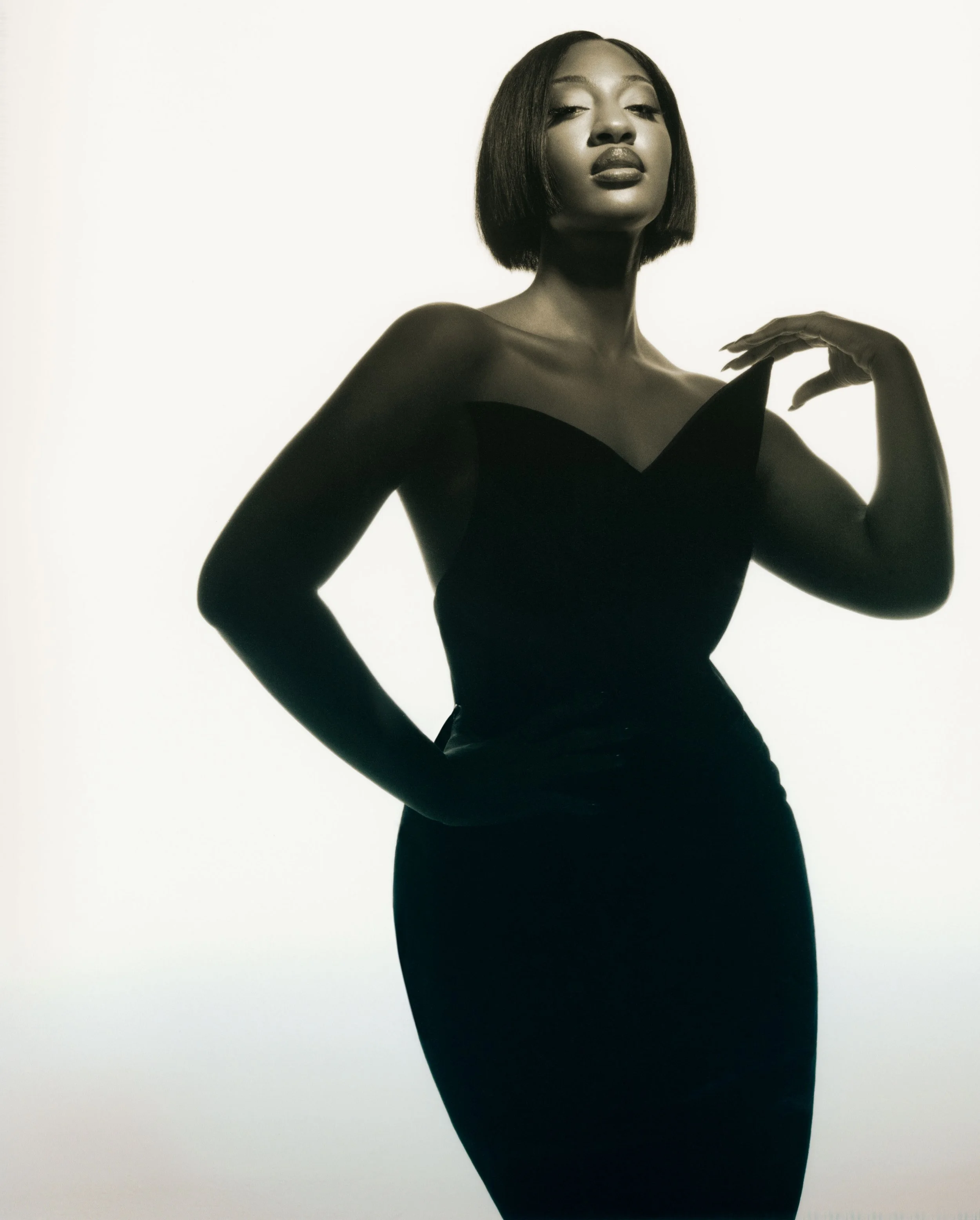 A black woman in a black dress with her left hand on her hip and right hand near her face, posing against a white background.