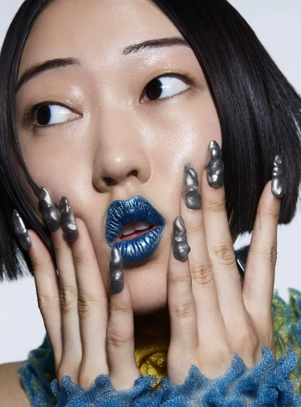 A close-up portrait of a woman with short black hair, blue metallic lipstick, and shiny blackened nails. She is touching her face with her fingers, creating an artistic and bold look.
