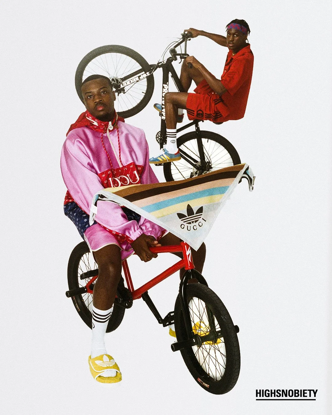 Two young men, one wearing a pink satin tracksuit and the other in a red outfit with a purple headband, pose with bicycles and a Gucci bag against a plain white background.