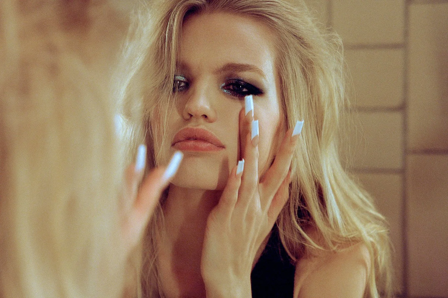 A woman with long blonde hair applying makeup to her eyes in front of a mirror.