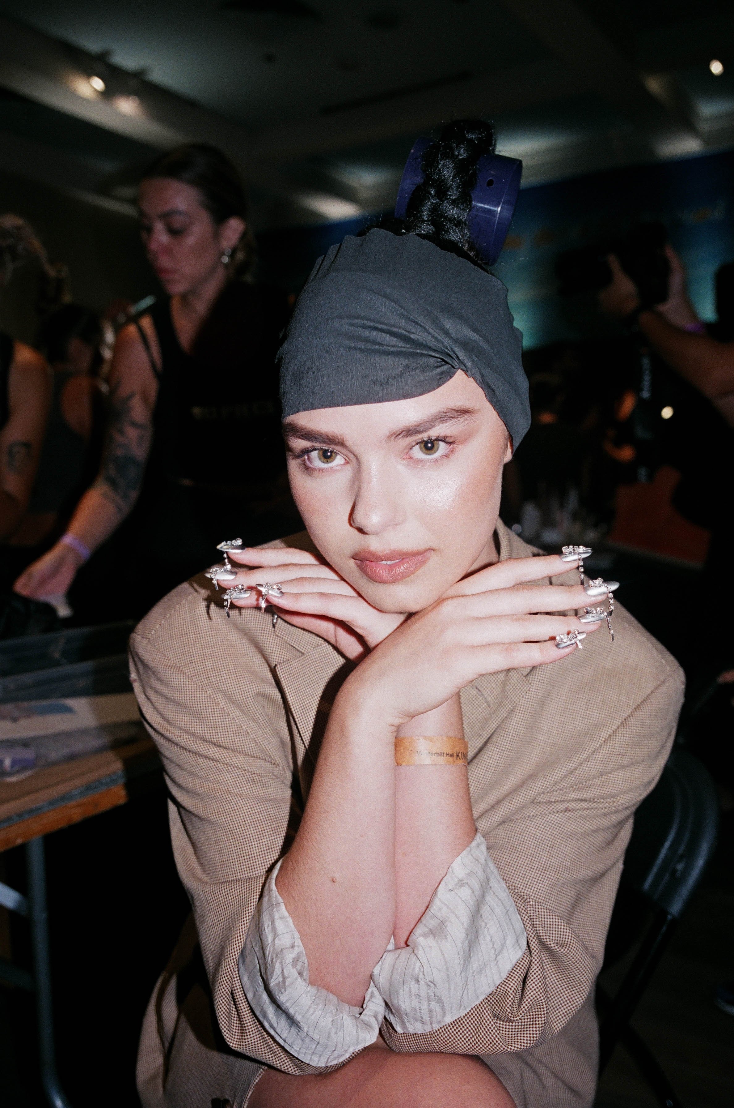 A woman with short brown hair, wearing a gray head scarf with a blue hair roller, posing with her hands under her chin in a dimly lit indoor event. She has multiple rings on her fingers and wears a beige blazer over a light shirt, with a wristband on her left wrist.