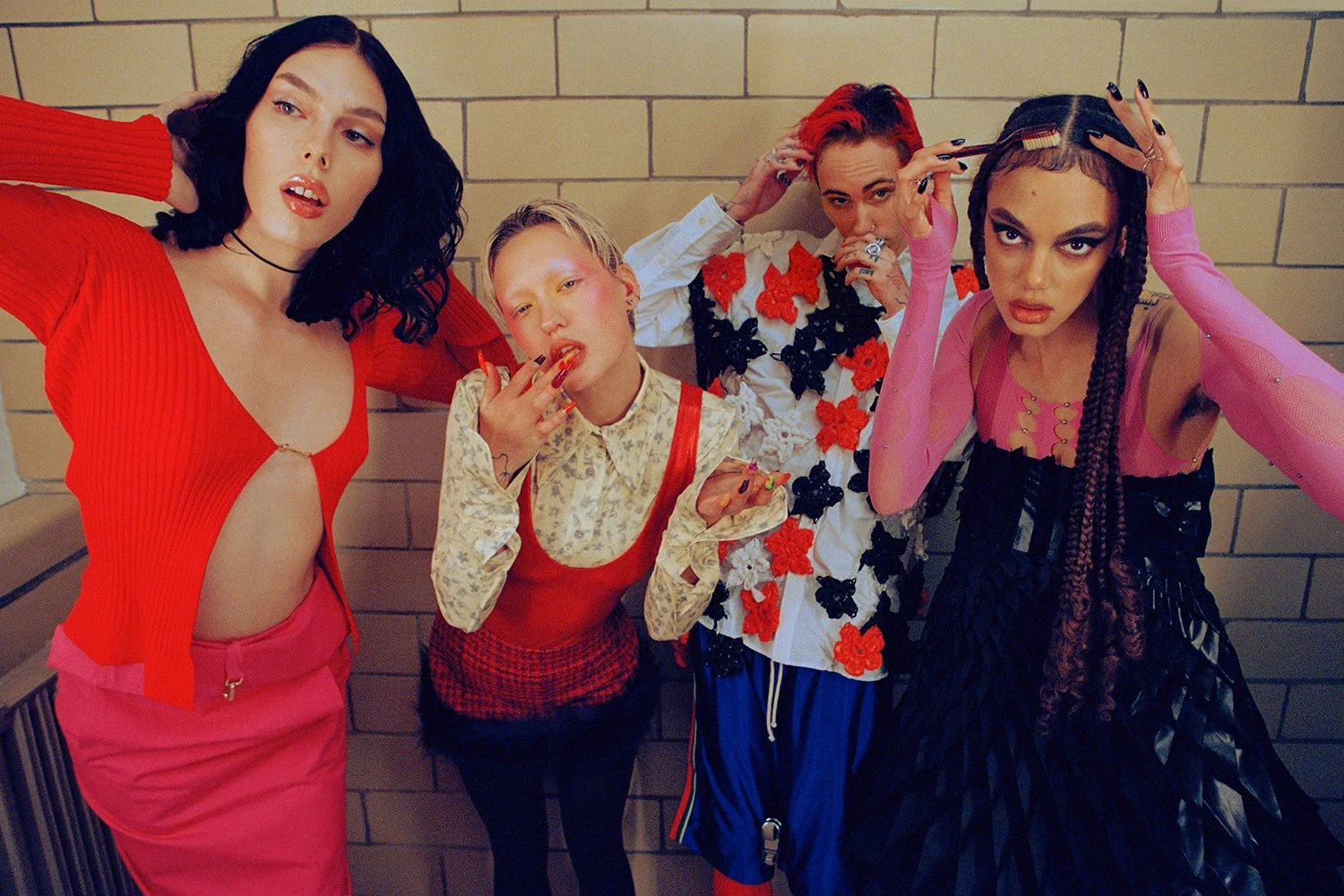 A group of five young women posing together against a brick wall, wearing colorful and fashionable clothing with bold makeup and hairstyles.