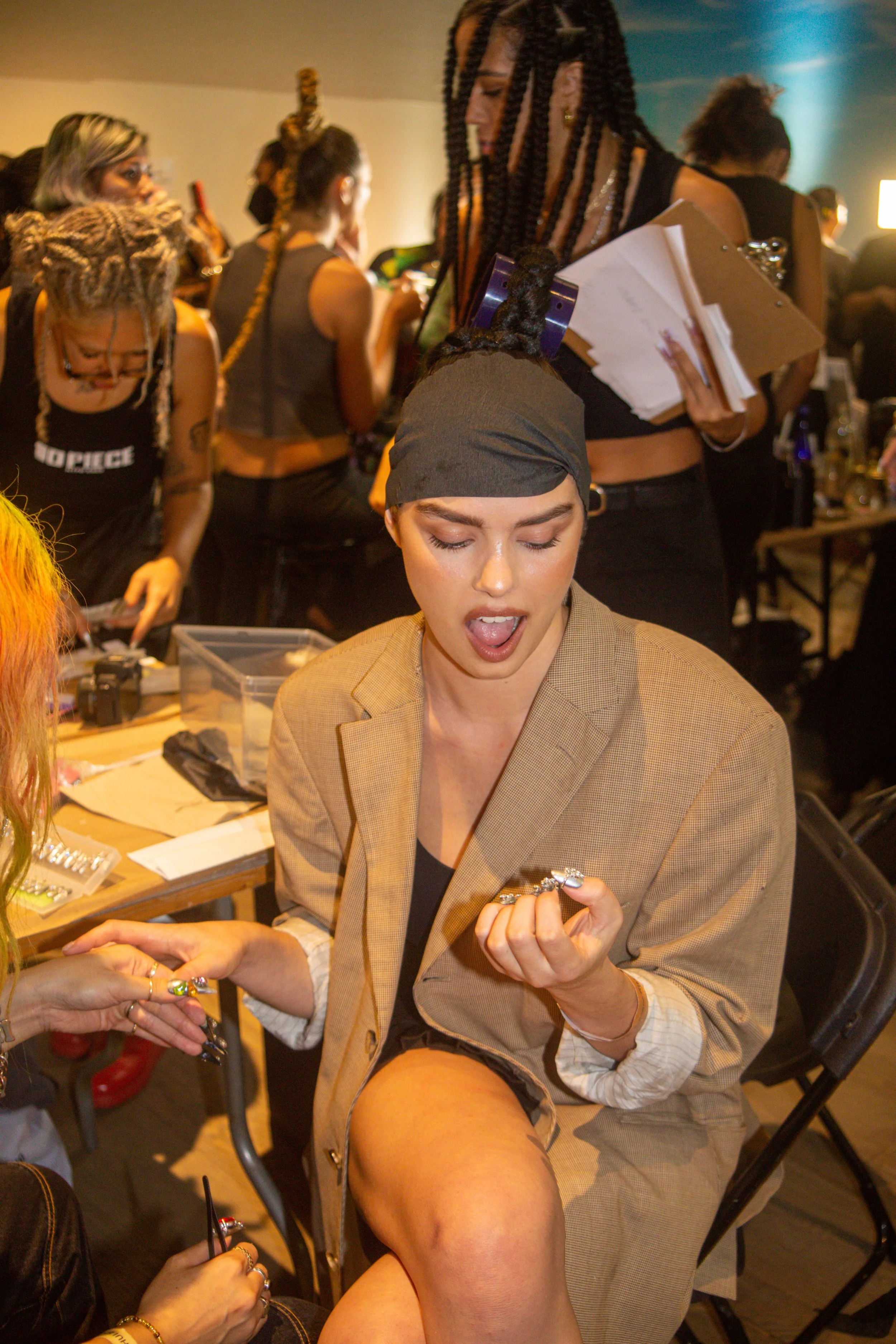 A young woman with braided hair, wearing a headscarf, beige blazer, and black top, is sitting at a table with her mouth open, examining rings in her hands, at a busy event where multiple people are working on jewelry.
