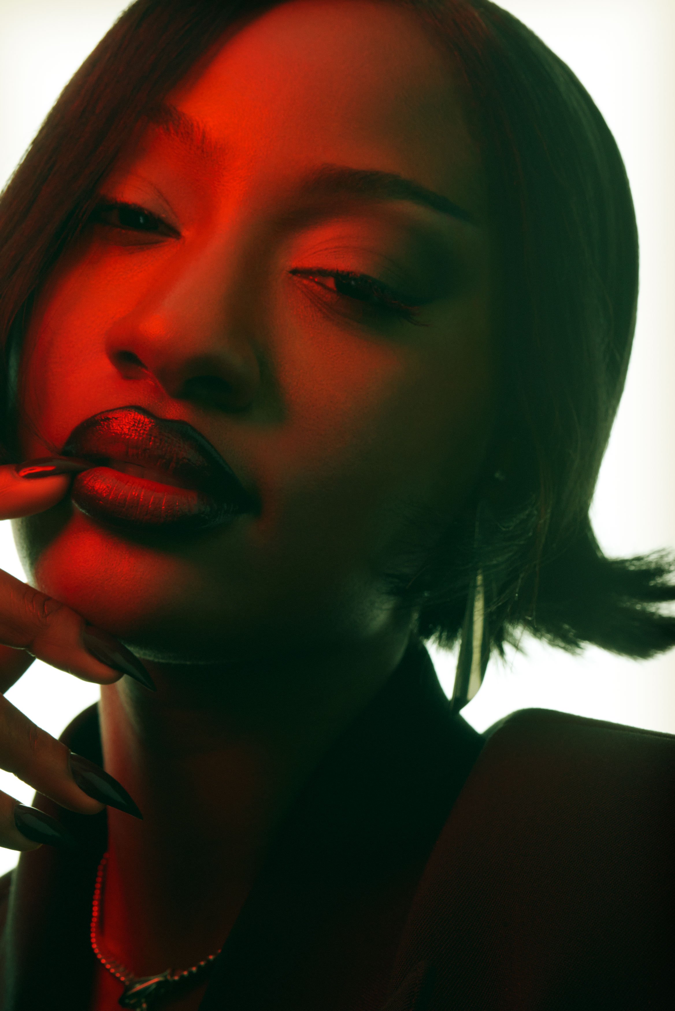 Close-up of a woman with dark hair, wearing dark lipstick and nails, posing with her hand near her face against a backlit background with red and green lighting.