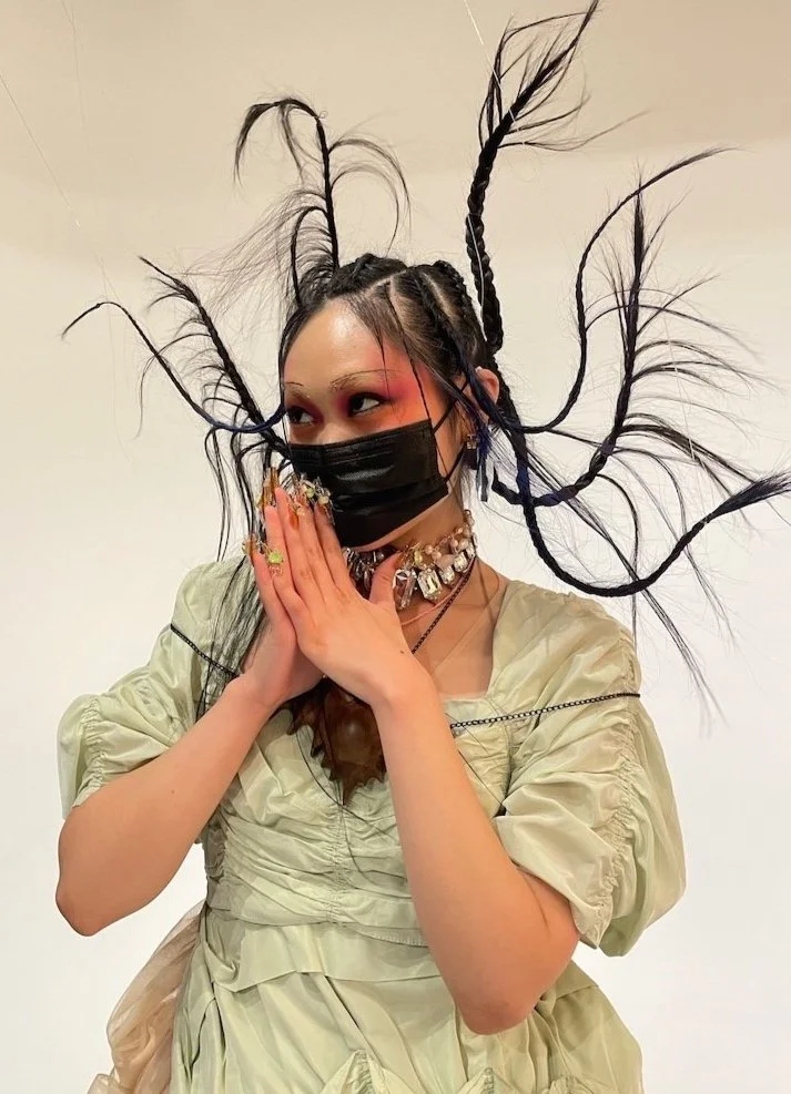 Person with braided and feathered hair, wearing a black face mask, jewelry, and a light-colored dress, posing with hands pressed together in front of their face.