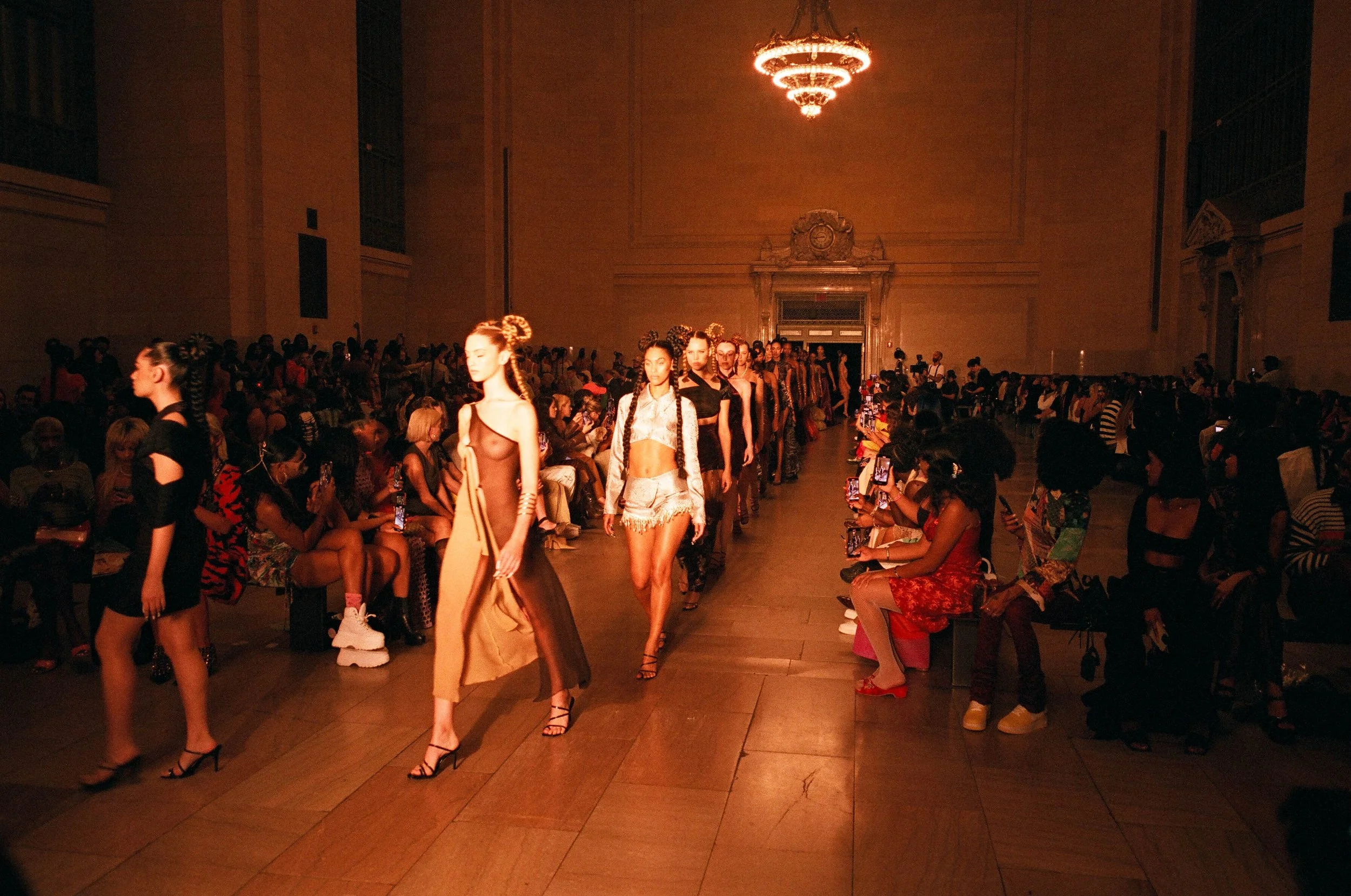 Models walk down runway during a fashion show in a large, elegant hall with a chandelier and high ceilings, audience seated on both sides of the runway taking photos.