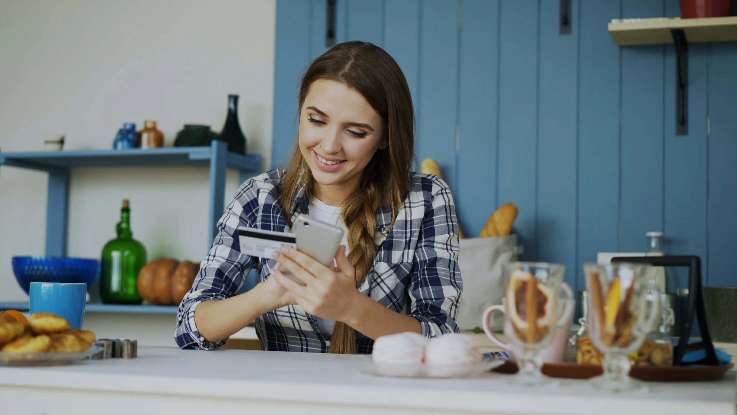 A young woman with long brown hair and a plaid shirt smiling while looking at her smartphone at a breakfast table with food and drinks in a cozy kitchen.