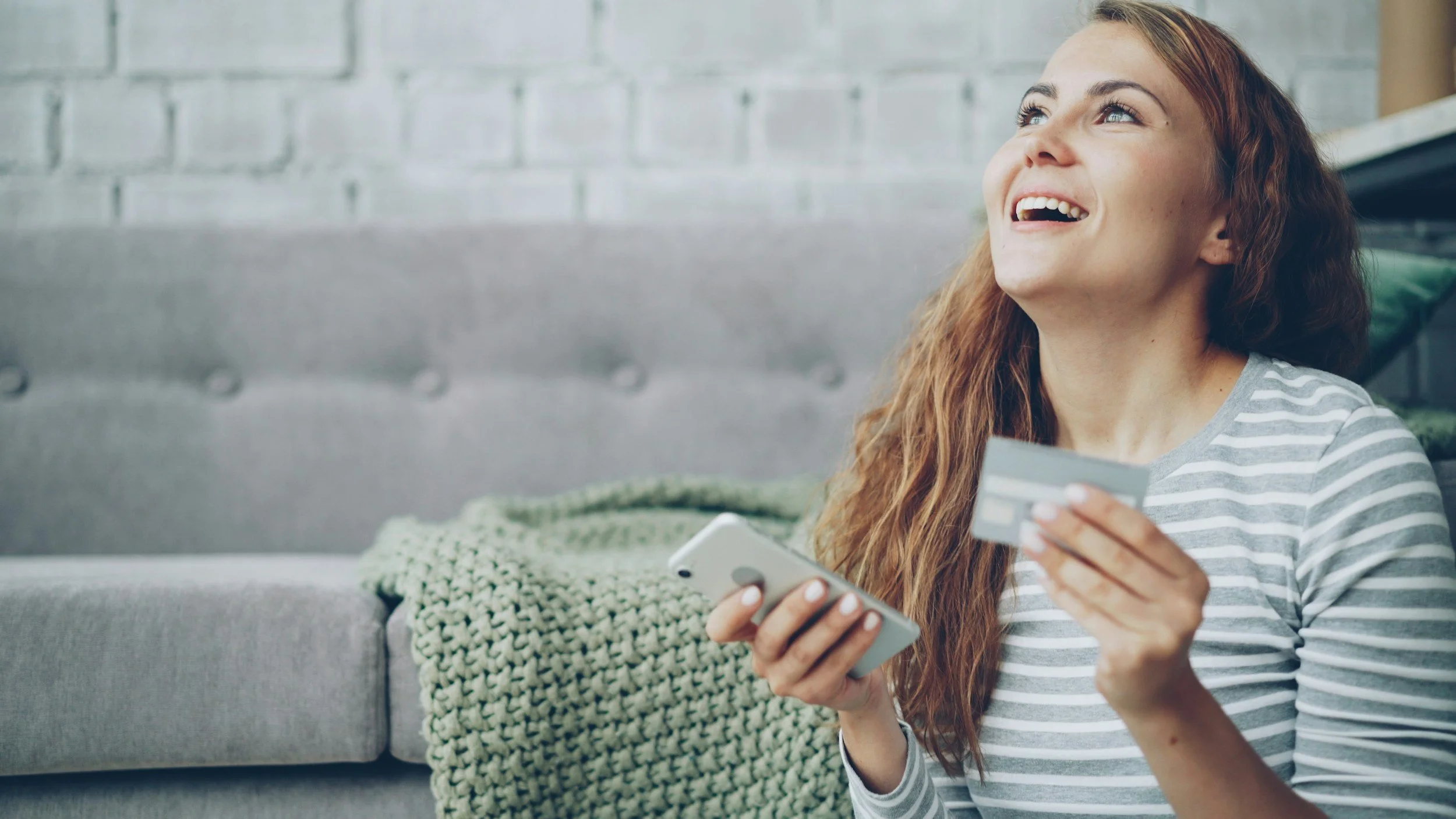 Woman with long red hair smiling while holding a smartphone and a credit card, sitting on a gray couch with a chunky knit blanket and a white brick wall background.
