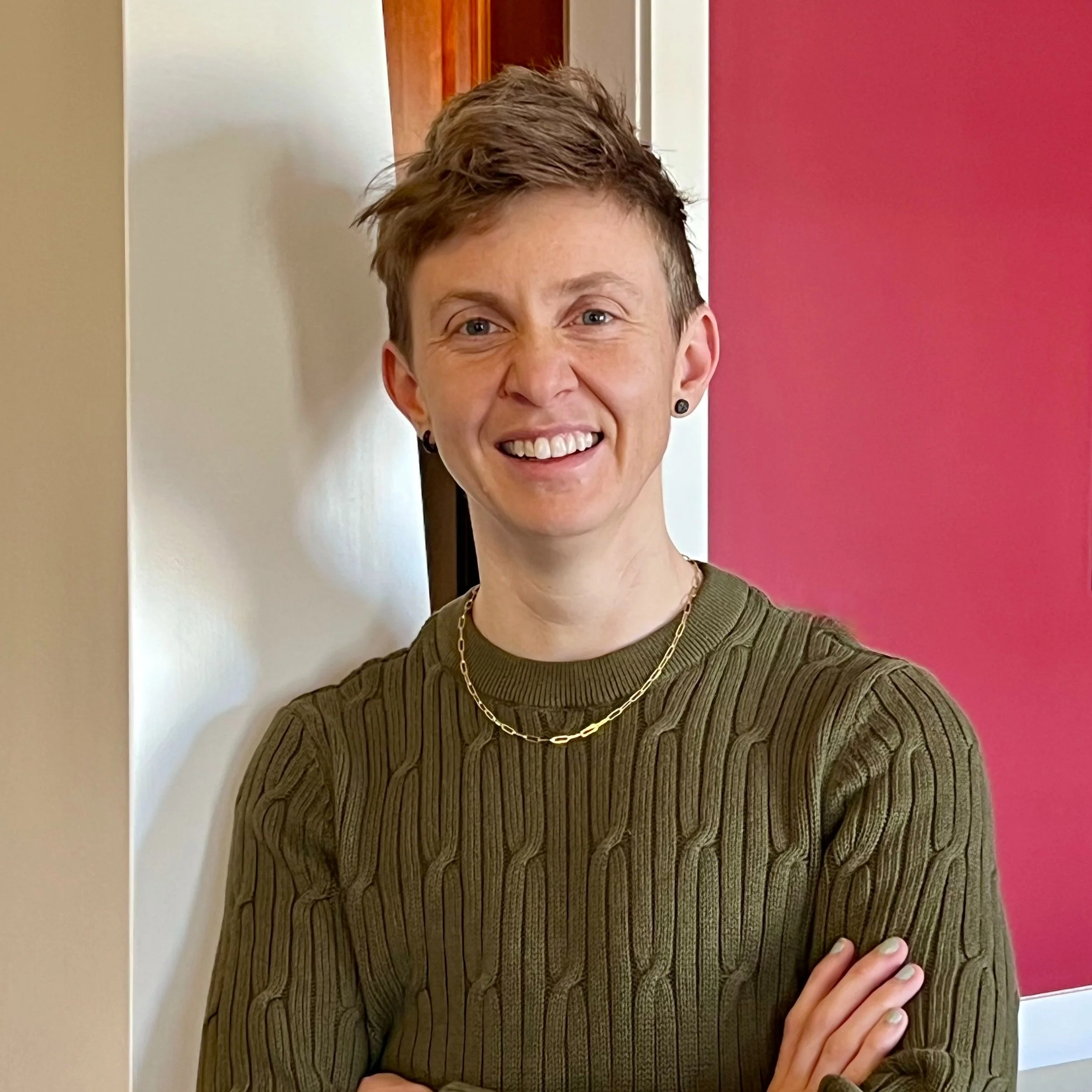 Close-up of a person with short, styled hair, smiling, wearing a green striped sweater, gold chain necklace, and black earrings, standing in front of a colorful wall with cream, brown, and pink sections.