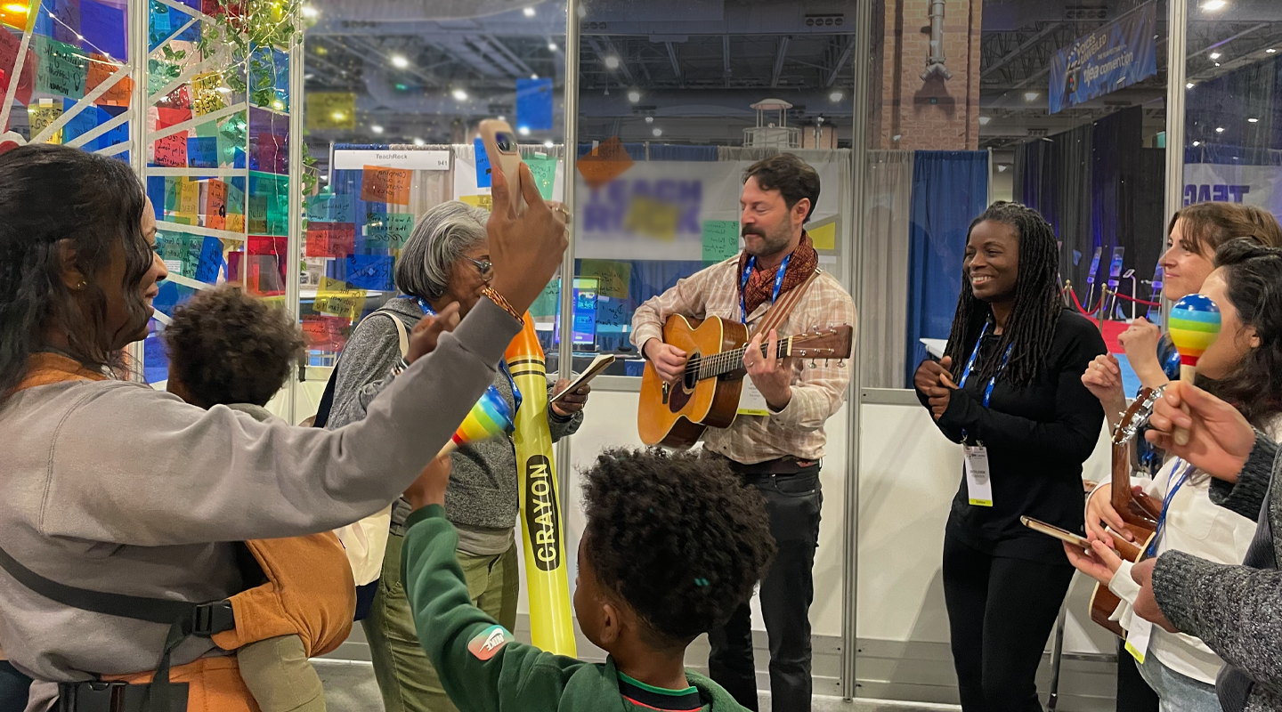 a diverse, smiling group of people stand in a circle inside a brightly colored booth, singing a song and playing guitar.