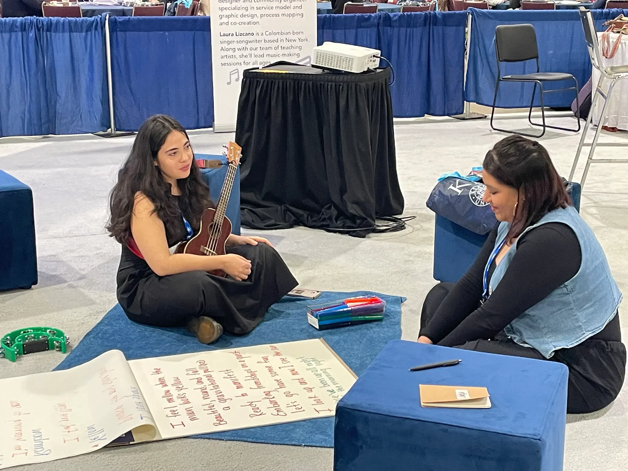 Two women sit on a blue mat with a ukulele and large lyrics sheet as they compose a song together.