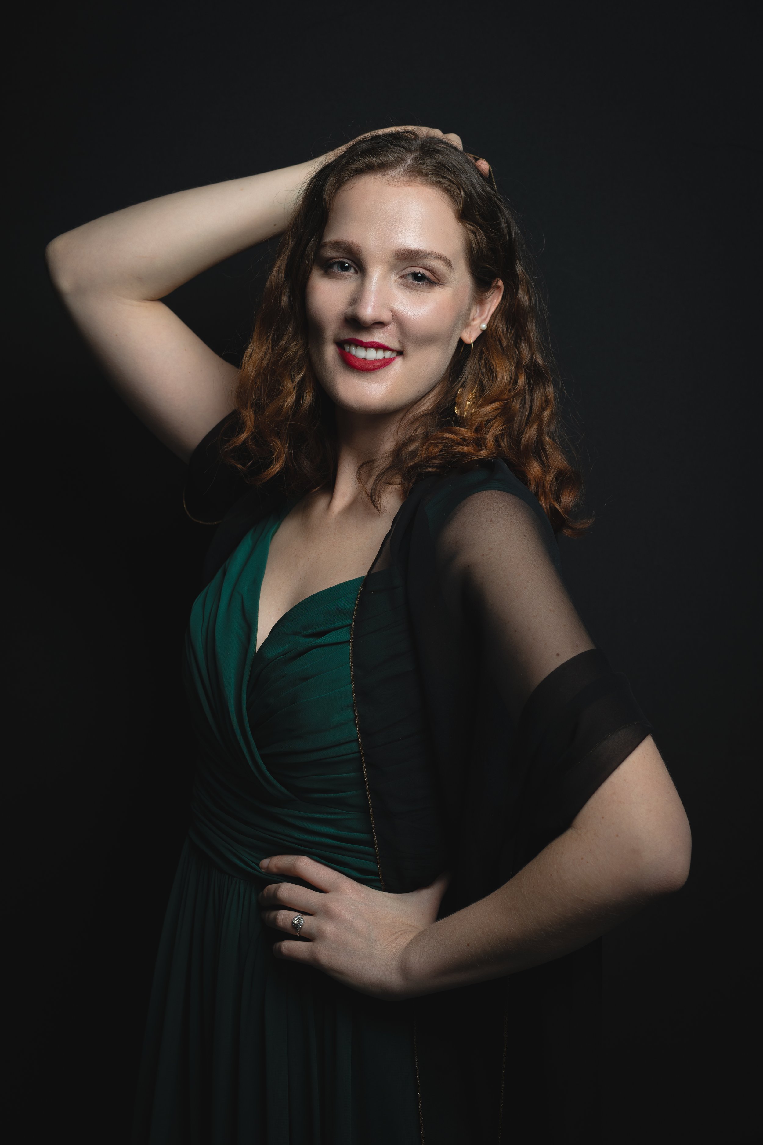 A woman with curly red hair wearing a green dress with sheer black sleeves, smiling and posing with her hand on her waist against a dark background.