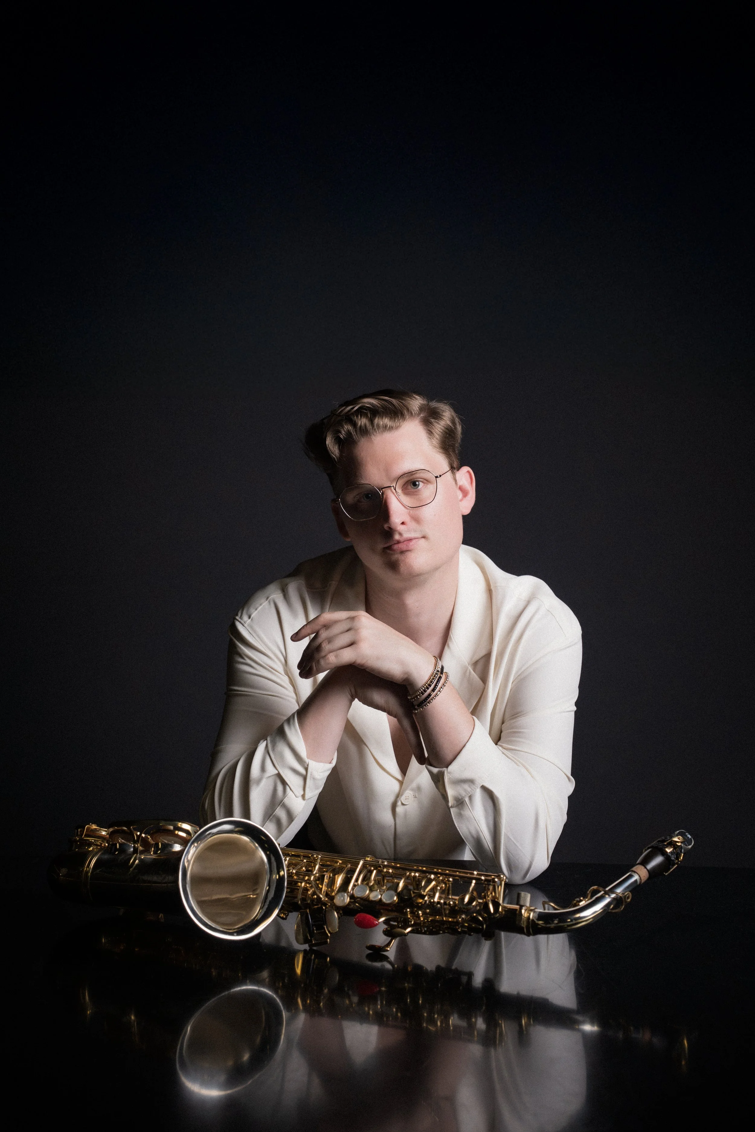 A man with glasses in a cream-colored shirt sits at a black table with a gold saxophone in front of him against a dark background.