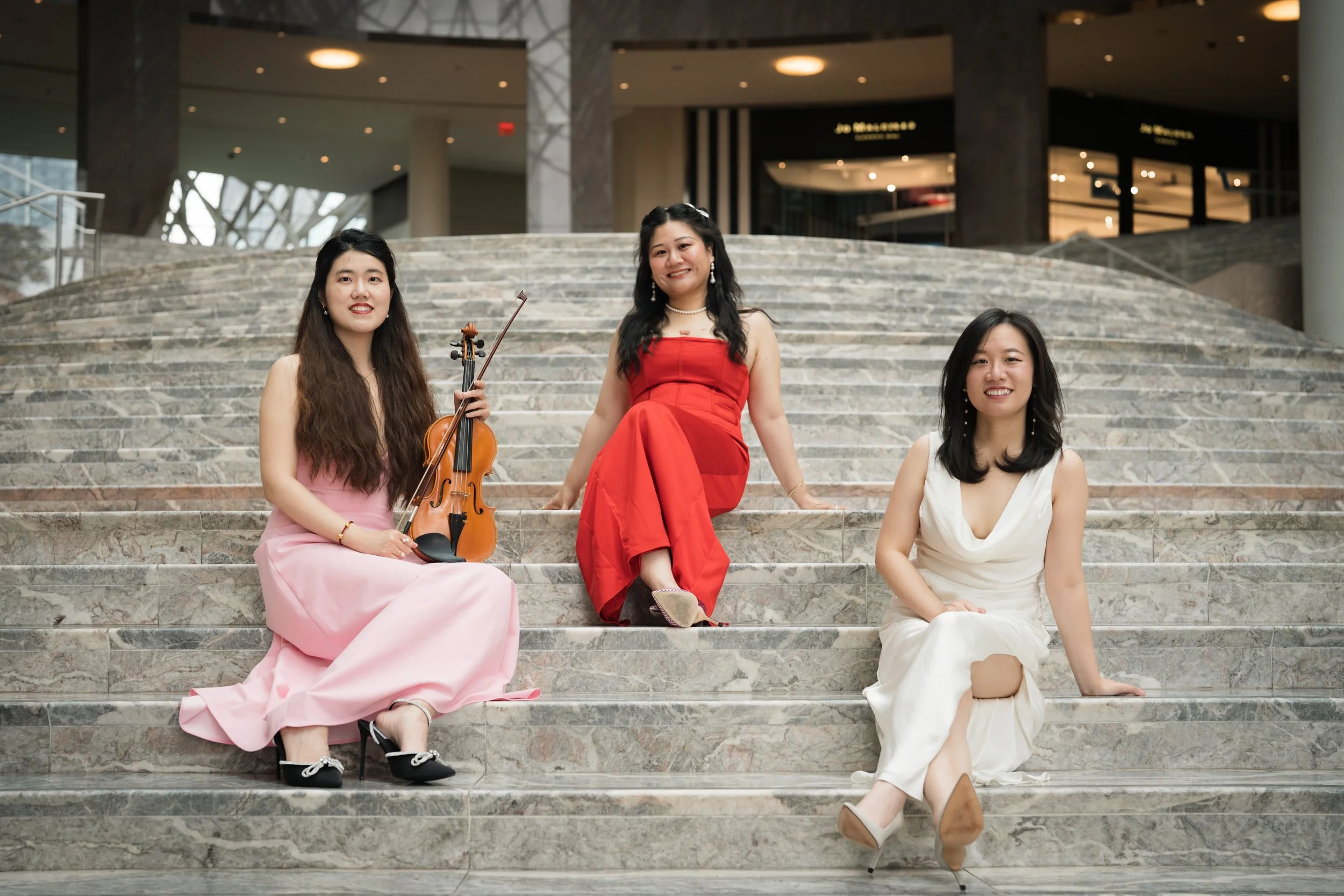Three women in formal dresses sitting on a staircase in a modern indoor setting, one holding a violin.