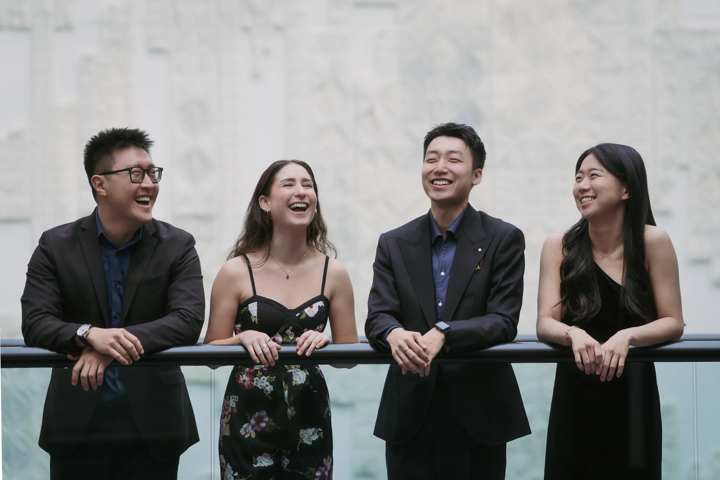 Four people dressed in formal attire leaning on a railing, laughing and talking together in an indoor setting.