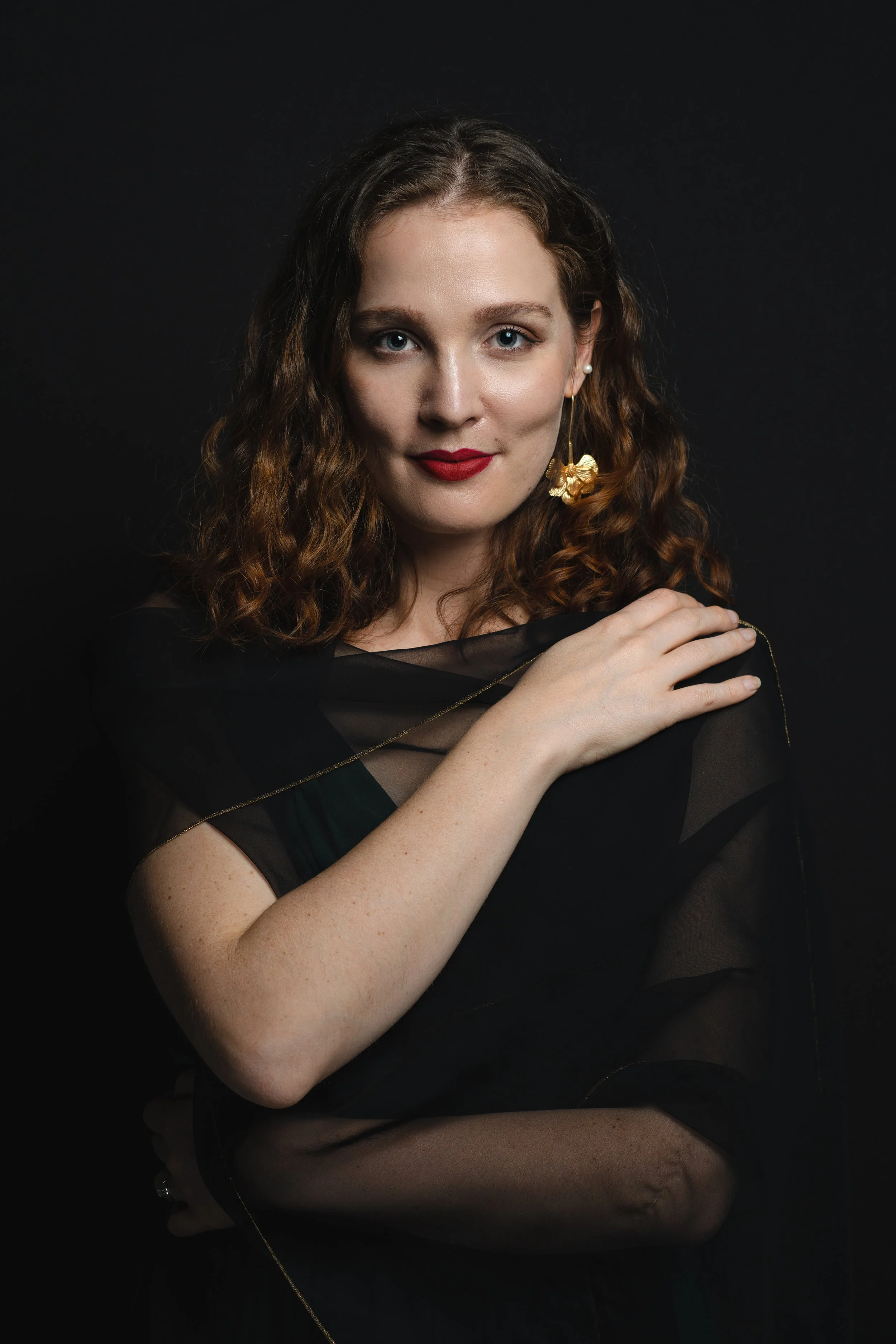 Portrait of a woman with curly red hair, wearing red lipstick, gold earrings, and a black sheer top, posing against a dark background.
