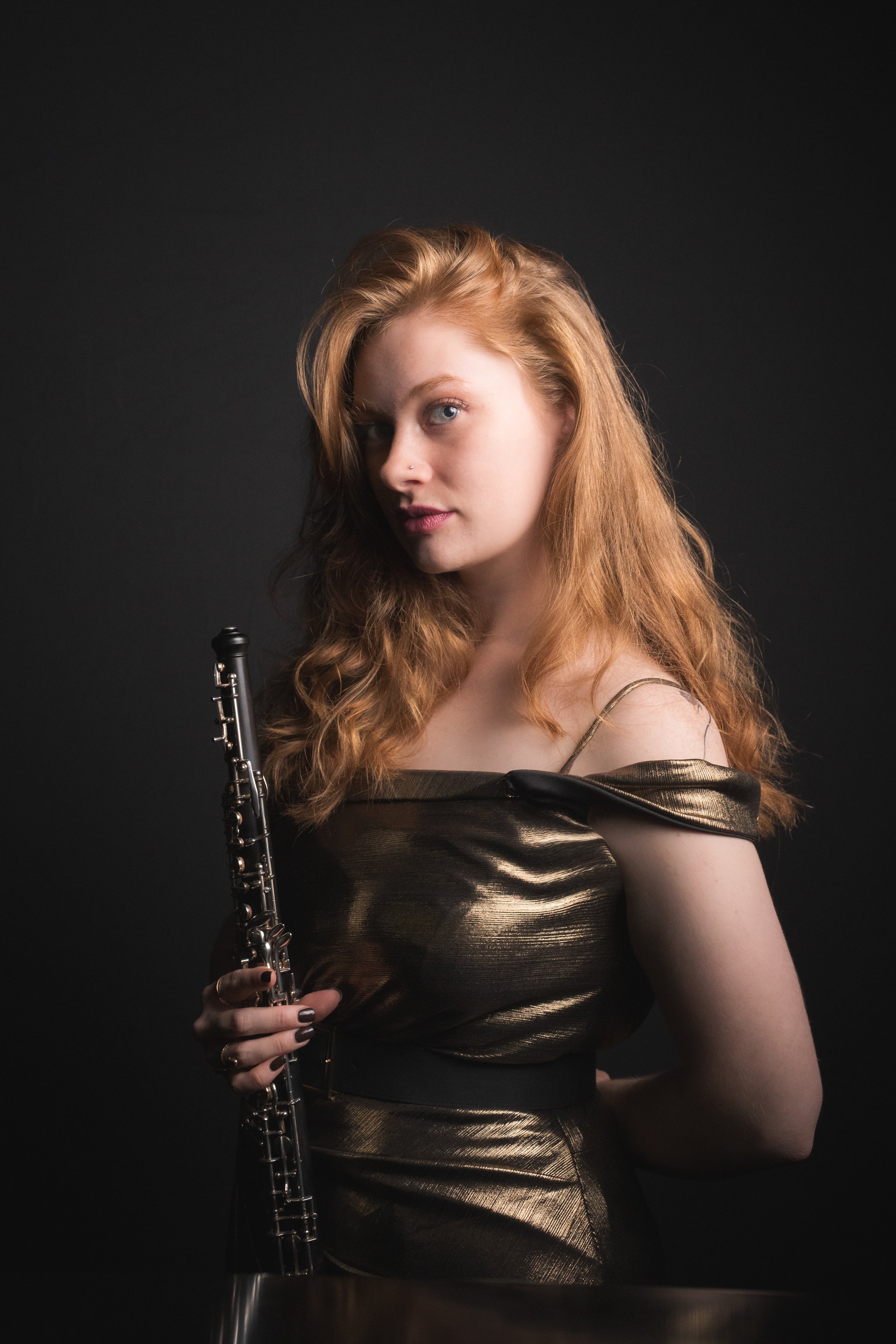 A young woman with long red hair, wearing a metallic gold off-shoulder dress, holding a clarinet against a black background.
