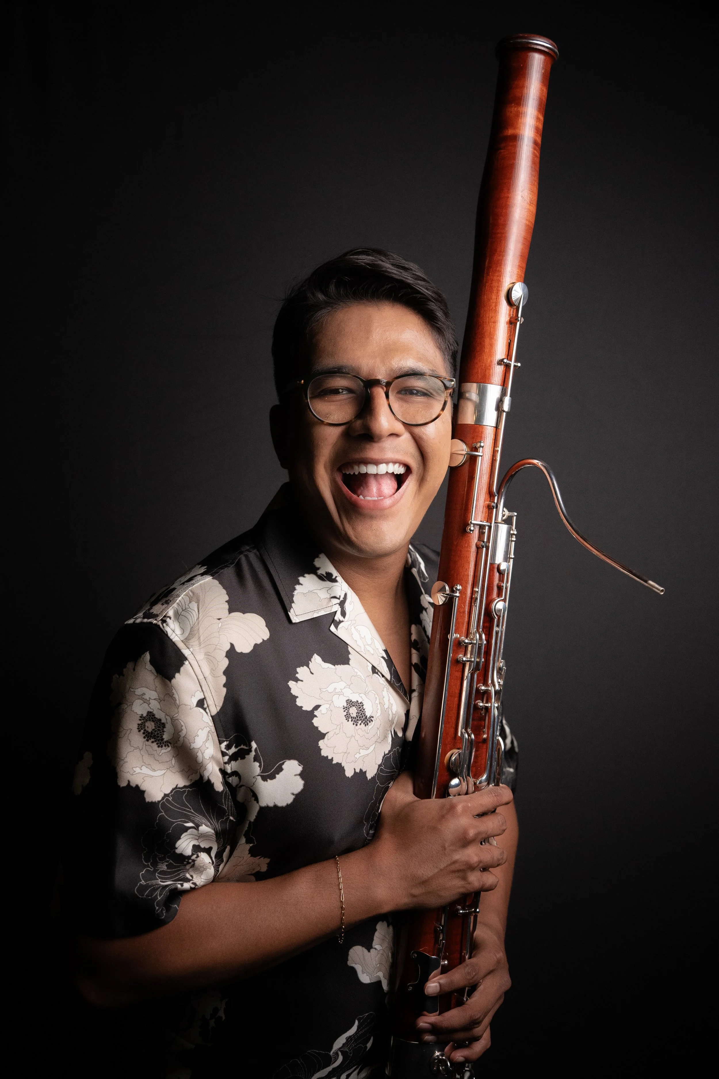 A smiling person holding a bassoon, wearing glasses and a floral shirt, against a dark background.