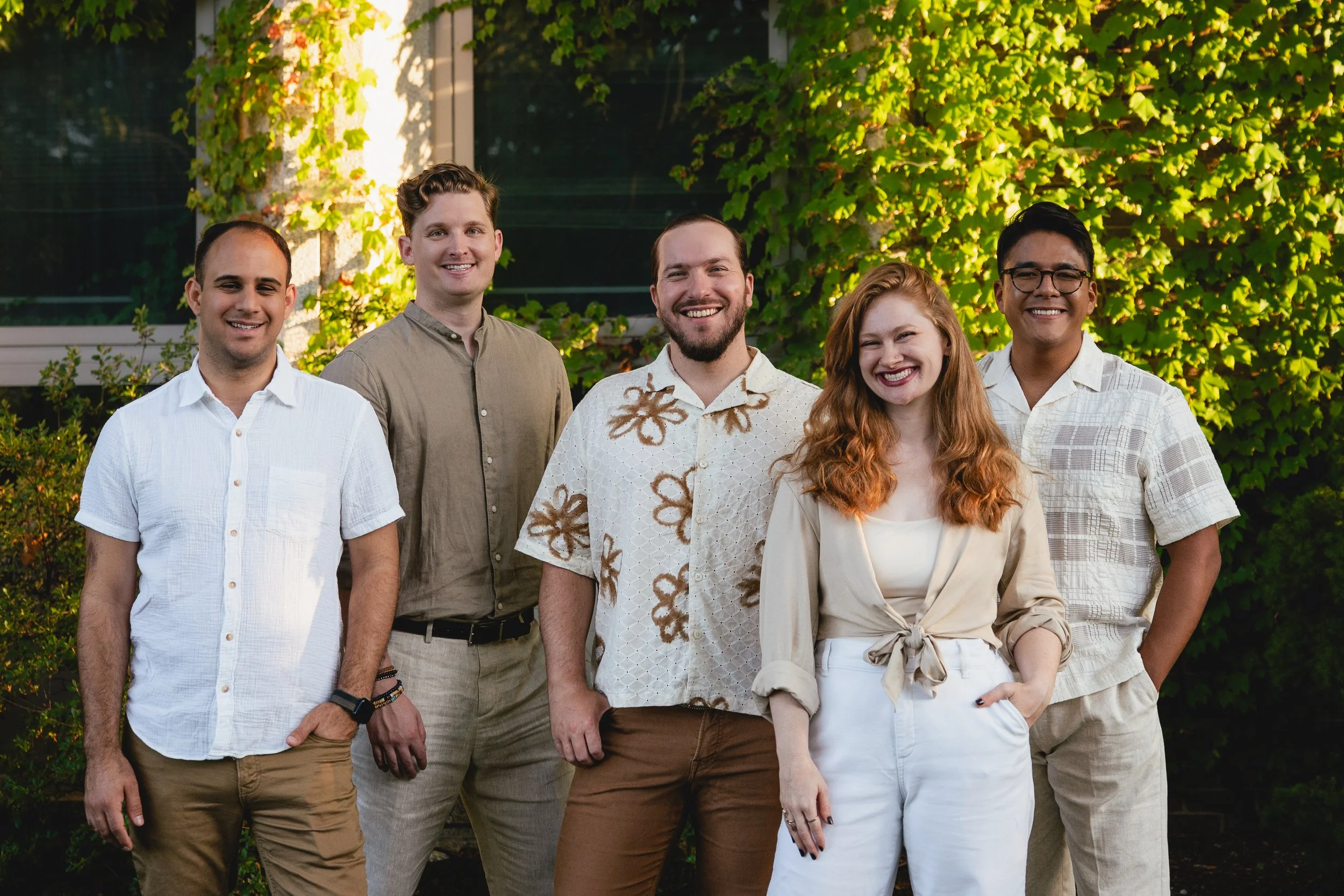 Group of six diverse young adults standing outdoors in front of green foliage, smiling at the camera.