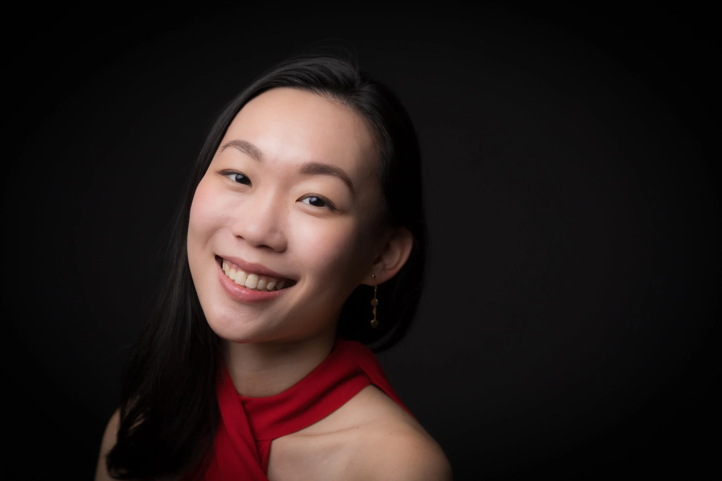 Portrait of a young woman with long dark hair, smiling with visible teeth, wearing a red sleeveless top, against a black background.