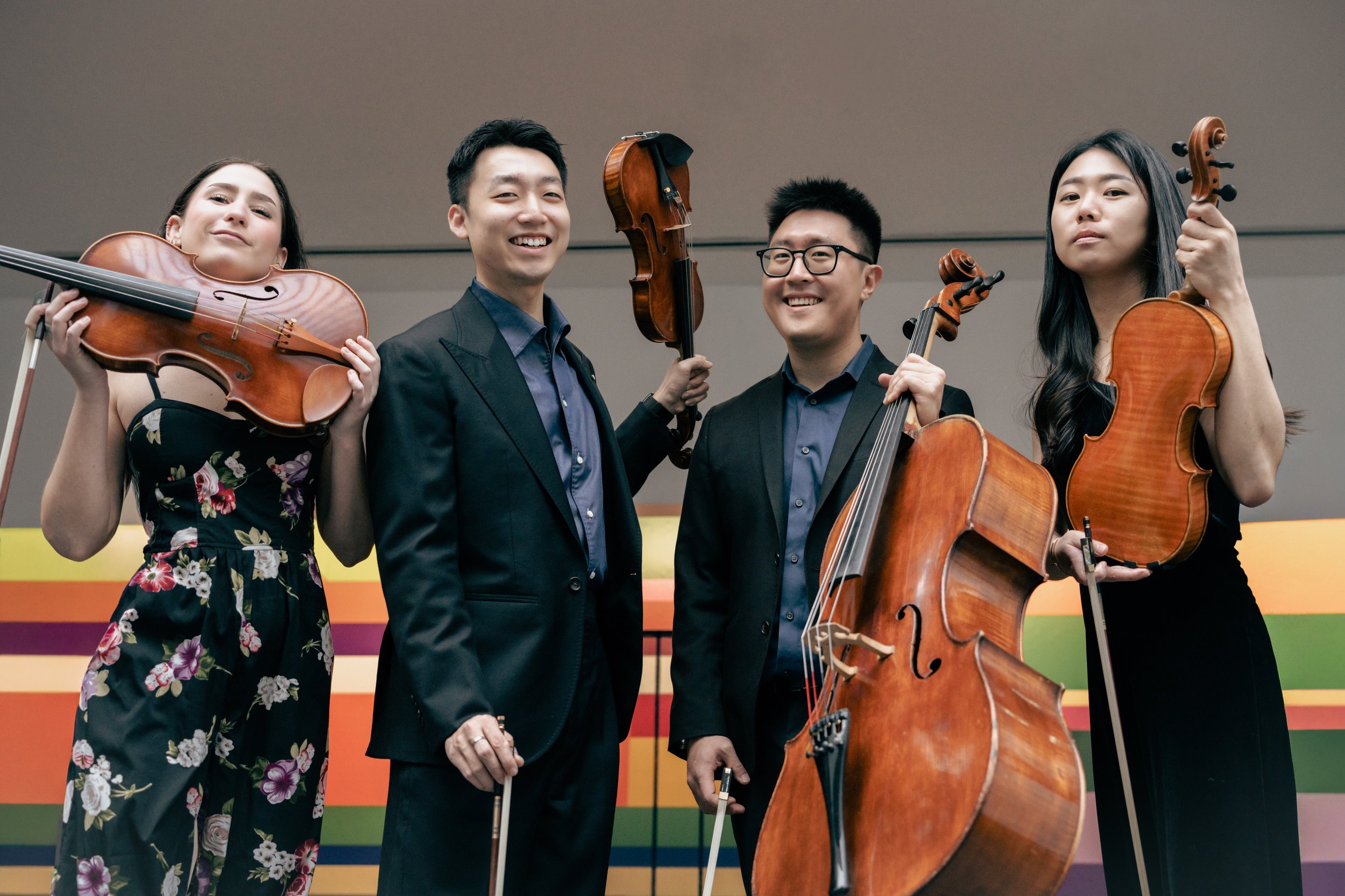 Four musicians holding string instruments, standing on stage with a colorful striped backdrop.