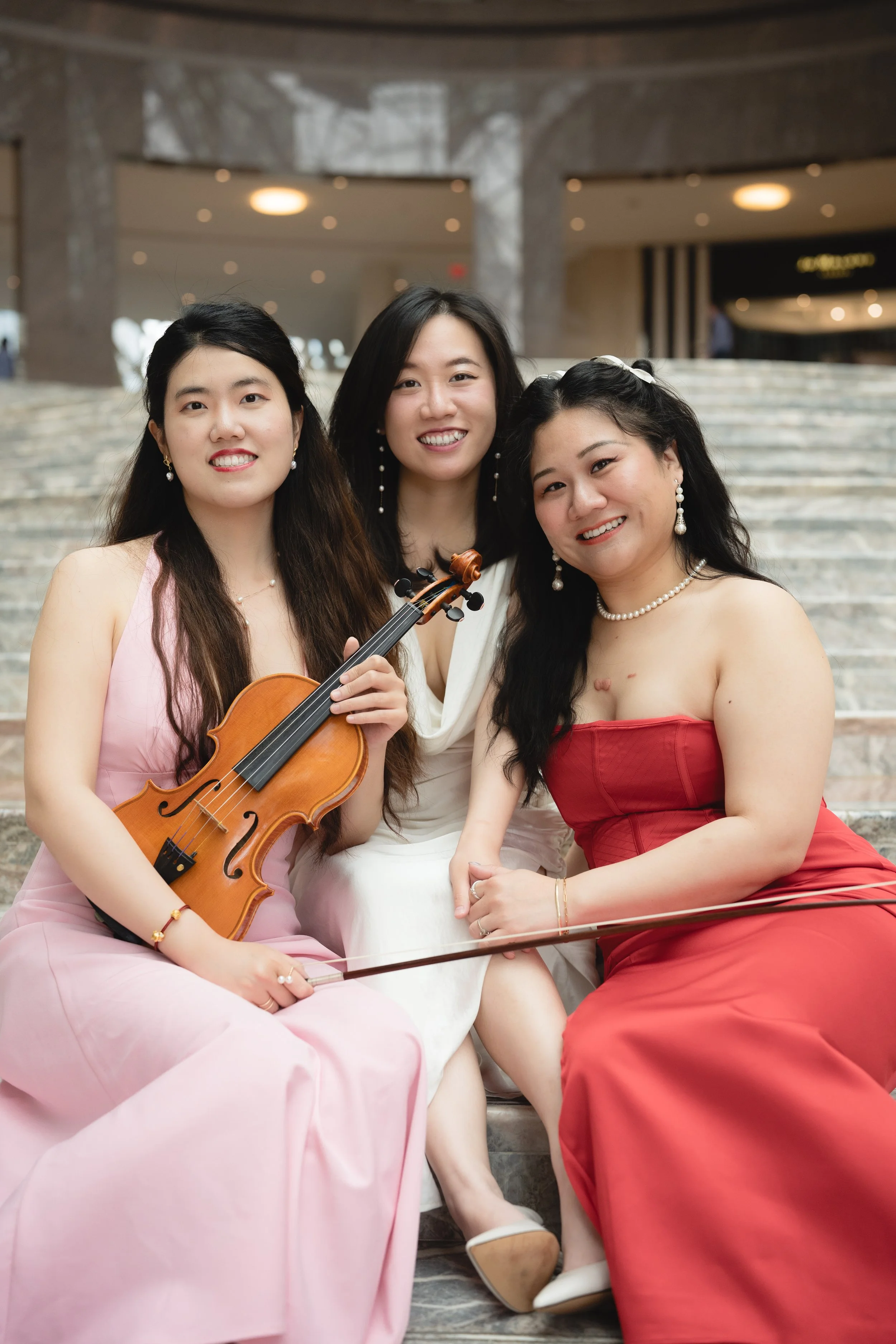 Three women sitting on steps indoors, one holding a violin, all dressed in evening gowns and smiling at the camera.