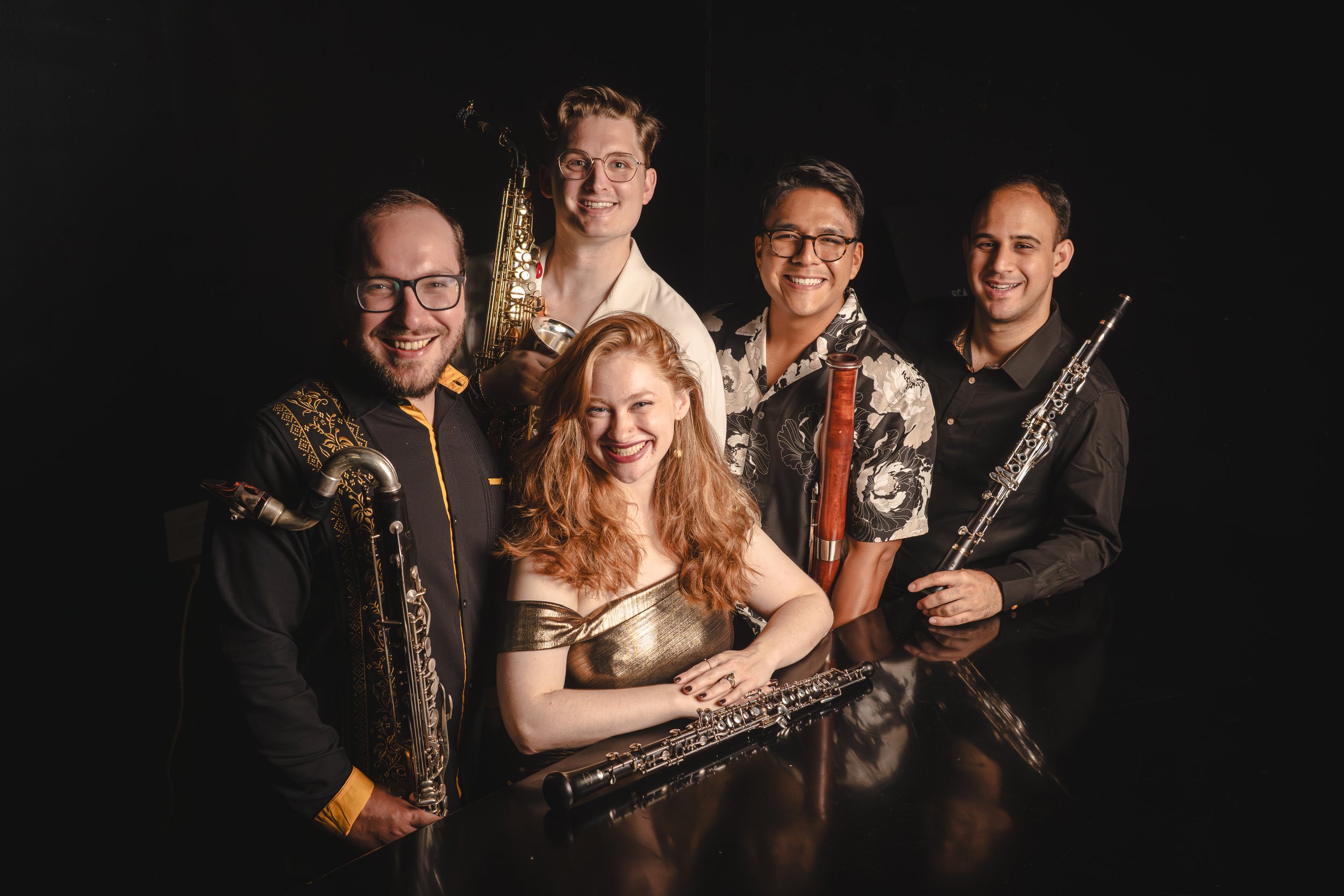 A group of six musicians, four men and one woman, posing with musical instruments against a black background. They are smiling and looking at the camera.