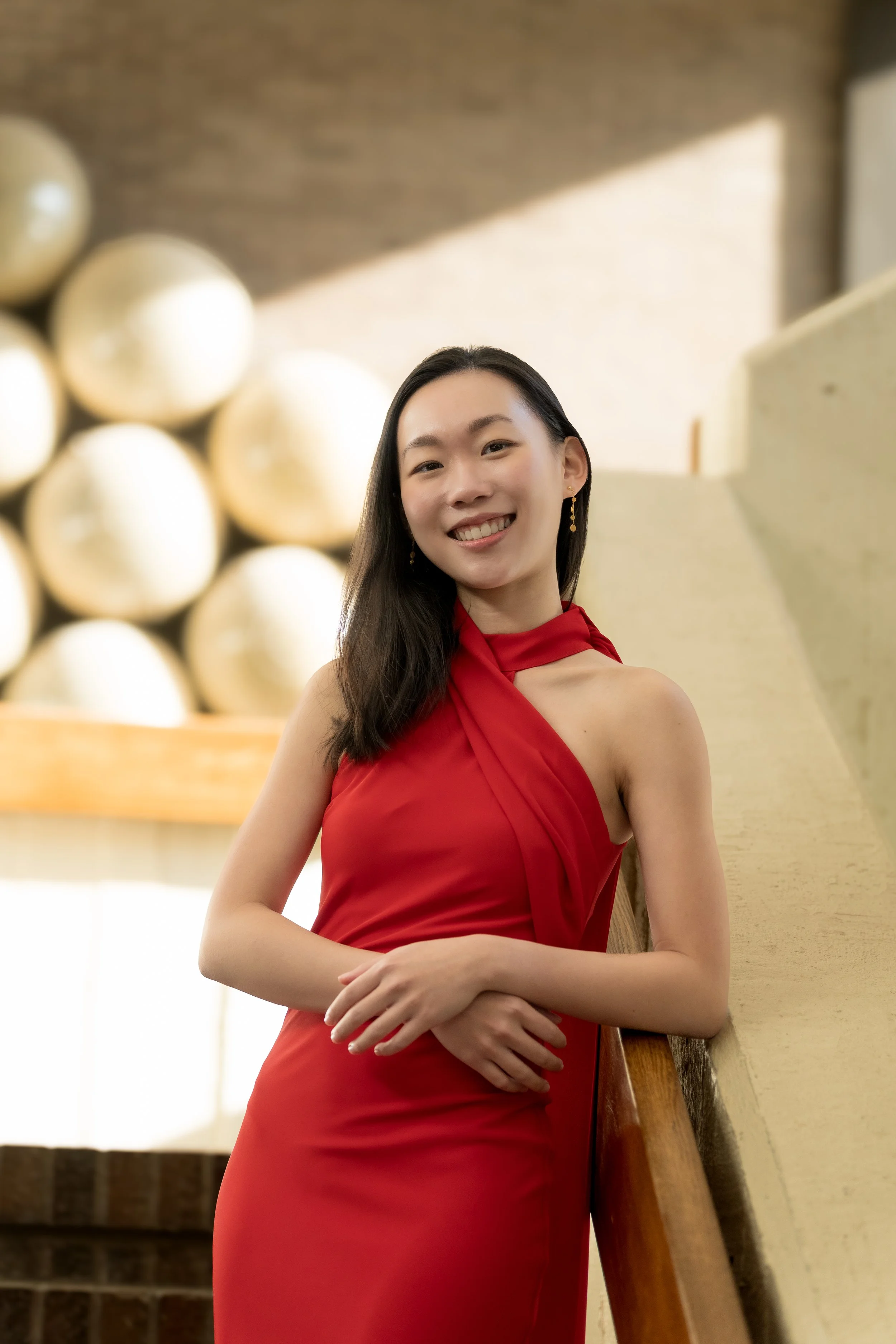 A woman in a red dress smiling and leaning against a railing in an indoor setting with a decorative wall of round objects in the background.