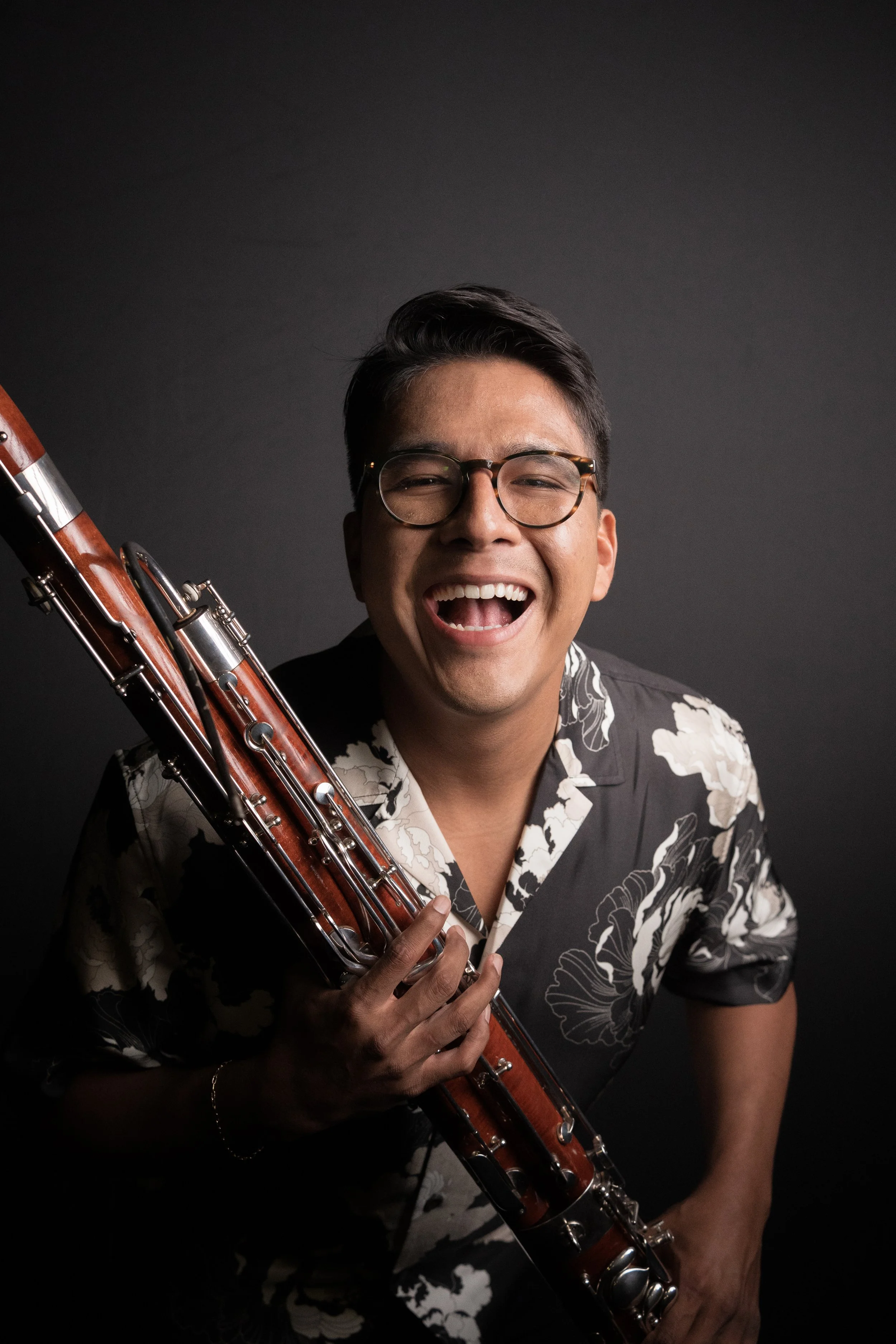 A man with glasses smiling and holding a bassoon against a dark background.