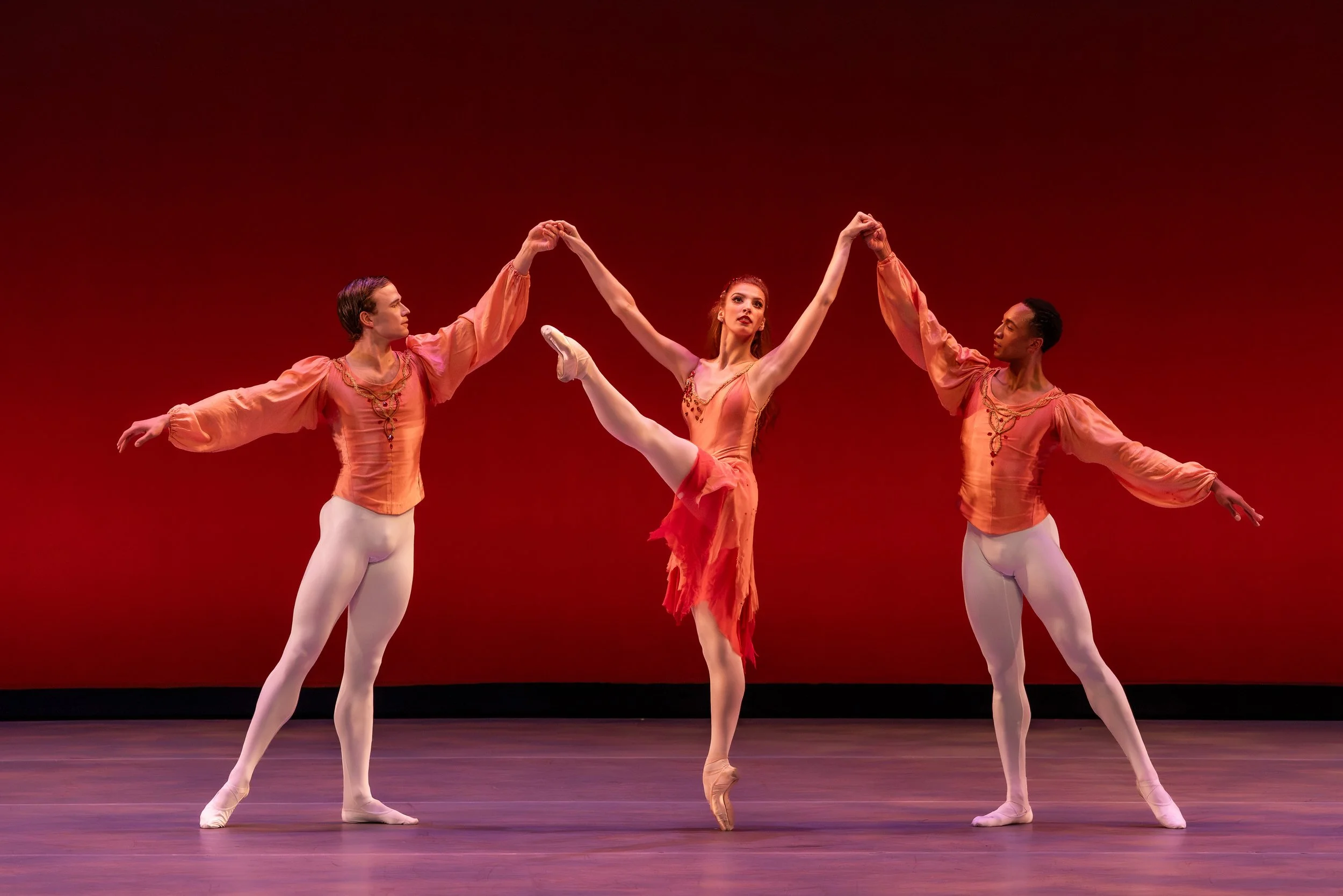 Three ballet dancers on stage, two supporting the central dancer in a ballet pose with one leg raised high and arms extended, against a red backdrop.