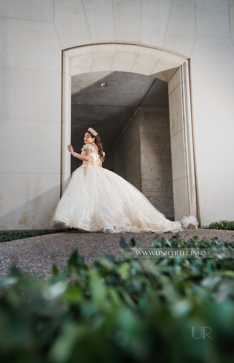 Portrait of a woman in an embellished white gown beside a stone arch, surrounded by greenery.