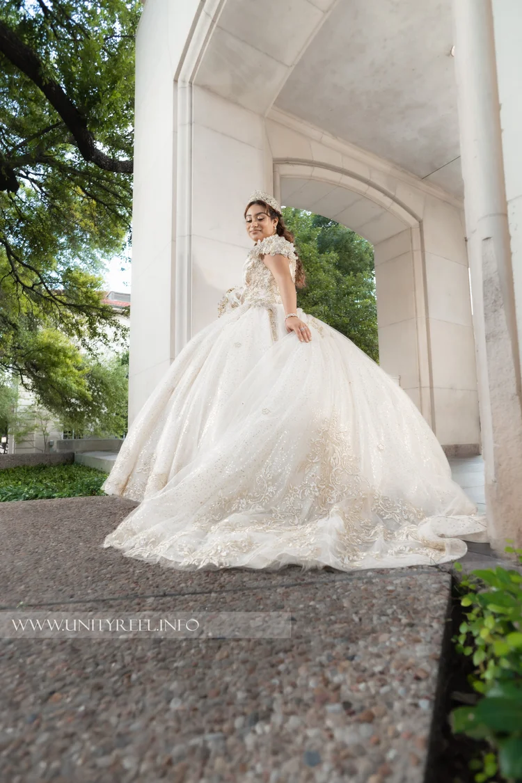 Portrait in an embroidered white gown beside stone archways and green trees.