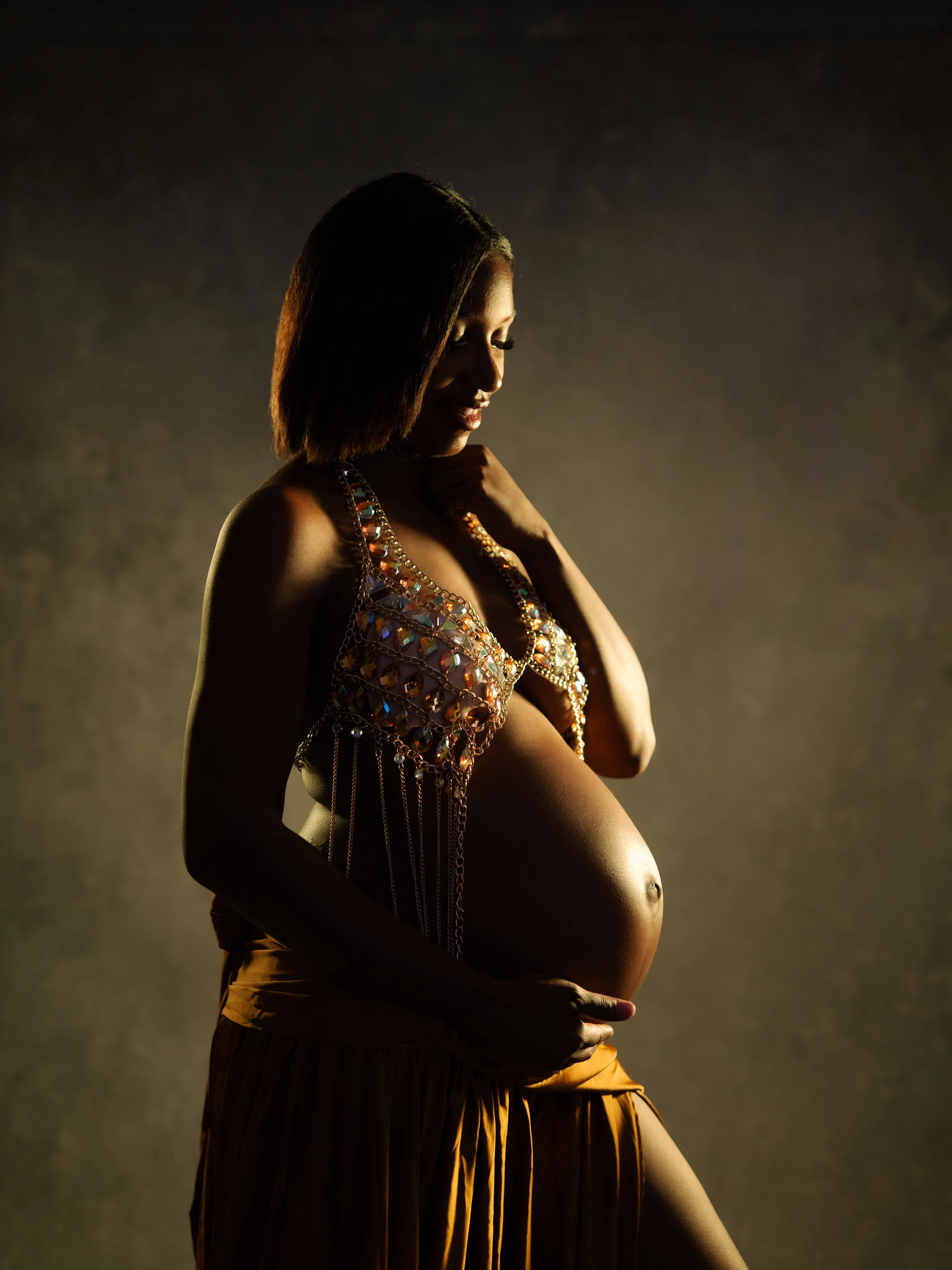 Pregnant woman with dark hair and makeup, wearing a beaded top and a flowy skirt, holding her belly, in profile, with low lighting and a dark background.