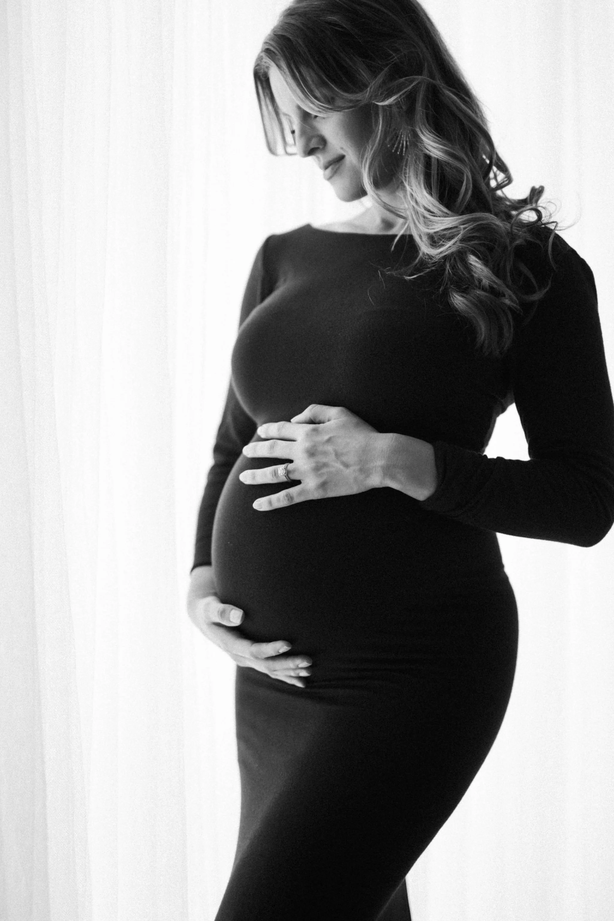Side view of a pregnant woman in a black dress, gently touching her abdomen with both hands, standing against a plain light-colored background.