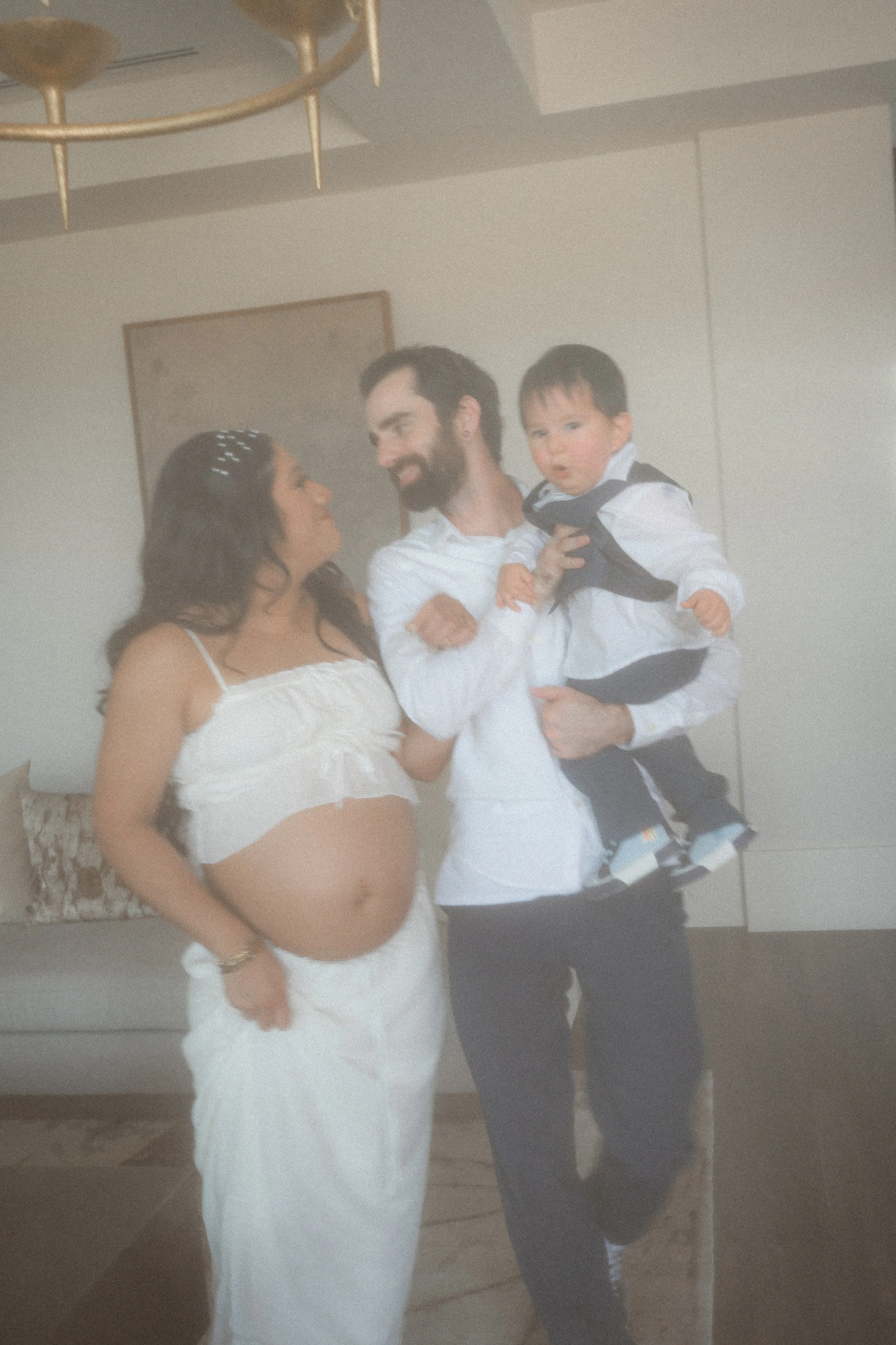 A pregnant woman in a white dress and bracelet, a man in a white shirt, and a young boy dressed in a white shirt, black vest, and black pants, are smiling and looking at each other in a room.