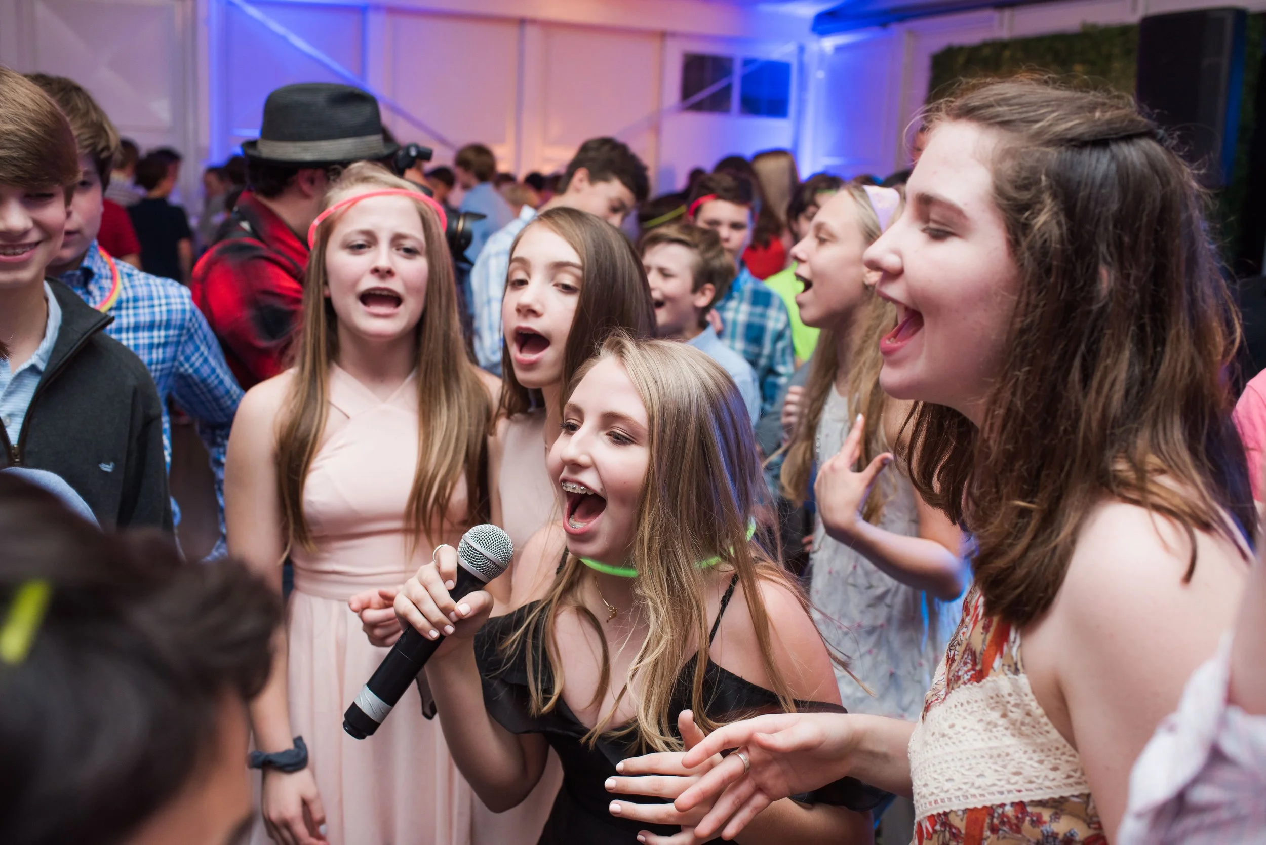 Group of young people at a party, some singing and holding a microphone, with colorful lighting in the background.