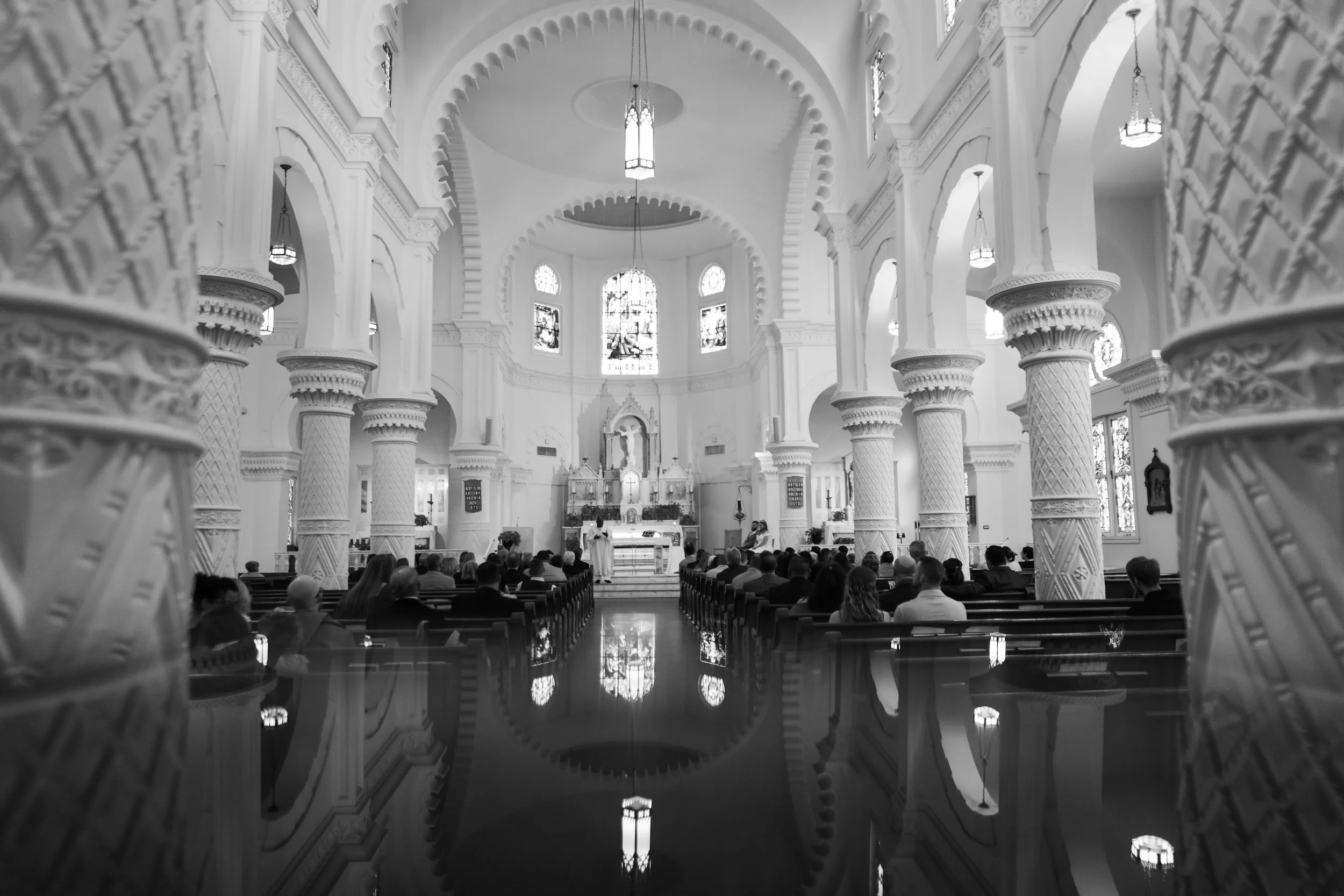 Interior of a church with ornate columns, chandeliers, stained glass windows, and a congregation seated during a service.