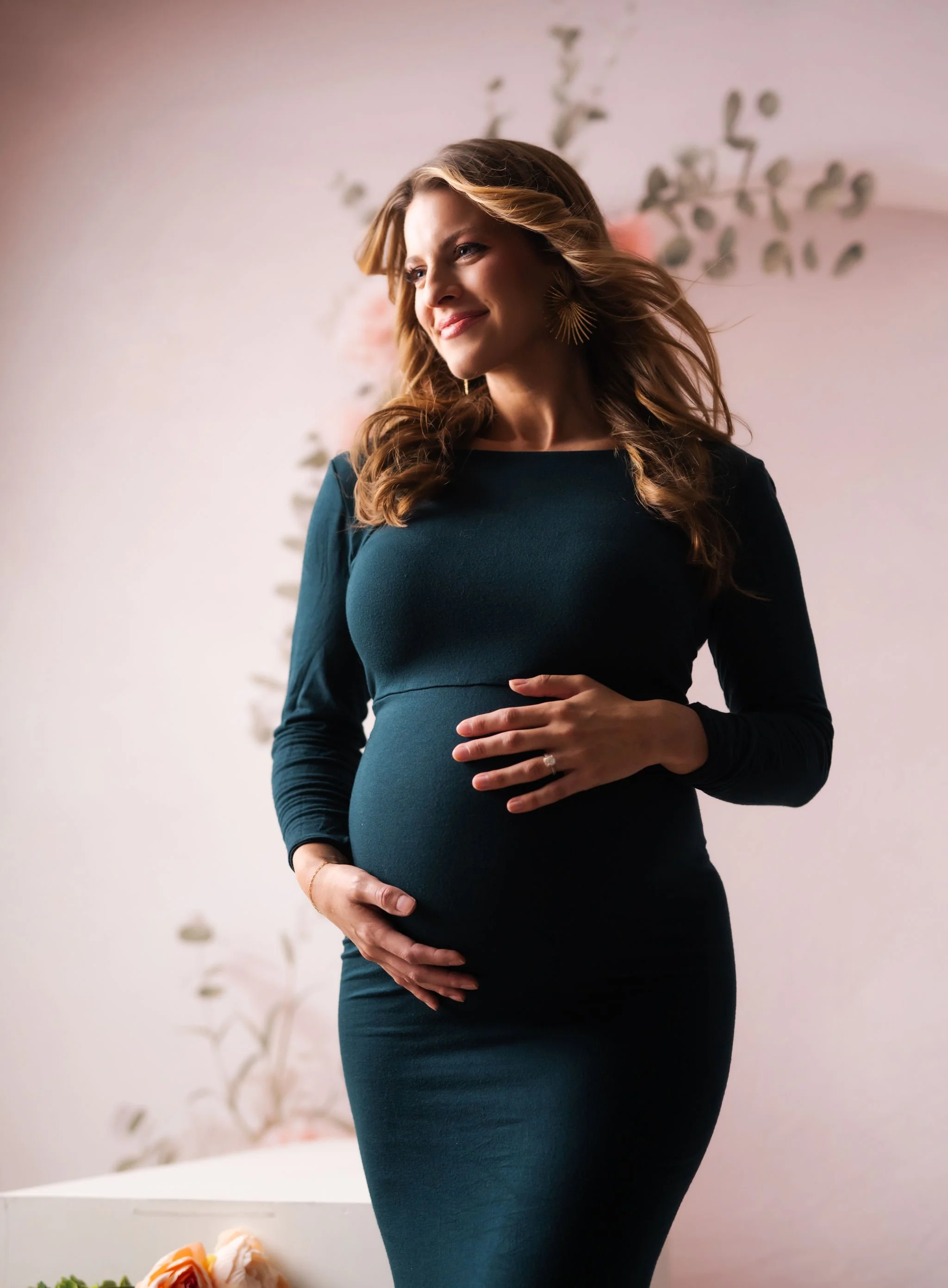 A pregnant woman with wavy brown hair and star-shaped earrings, smiling softly while holding her belly with both hands, standing in a softly lit room with a floral backdrop.