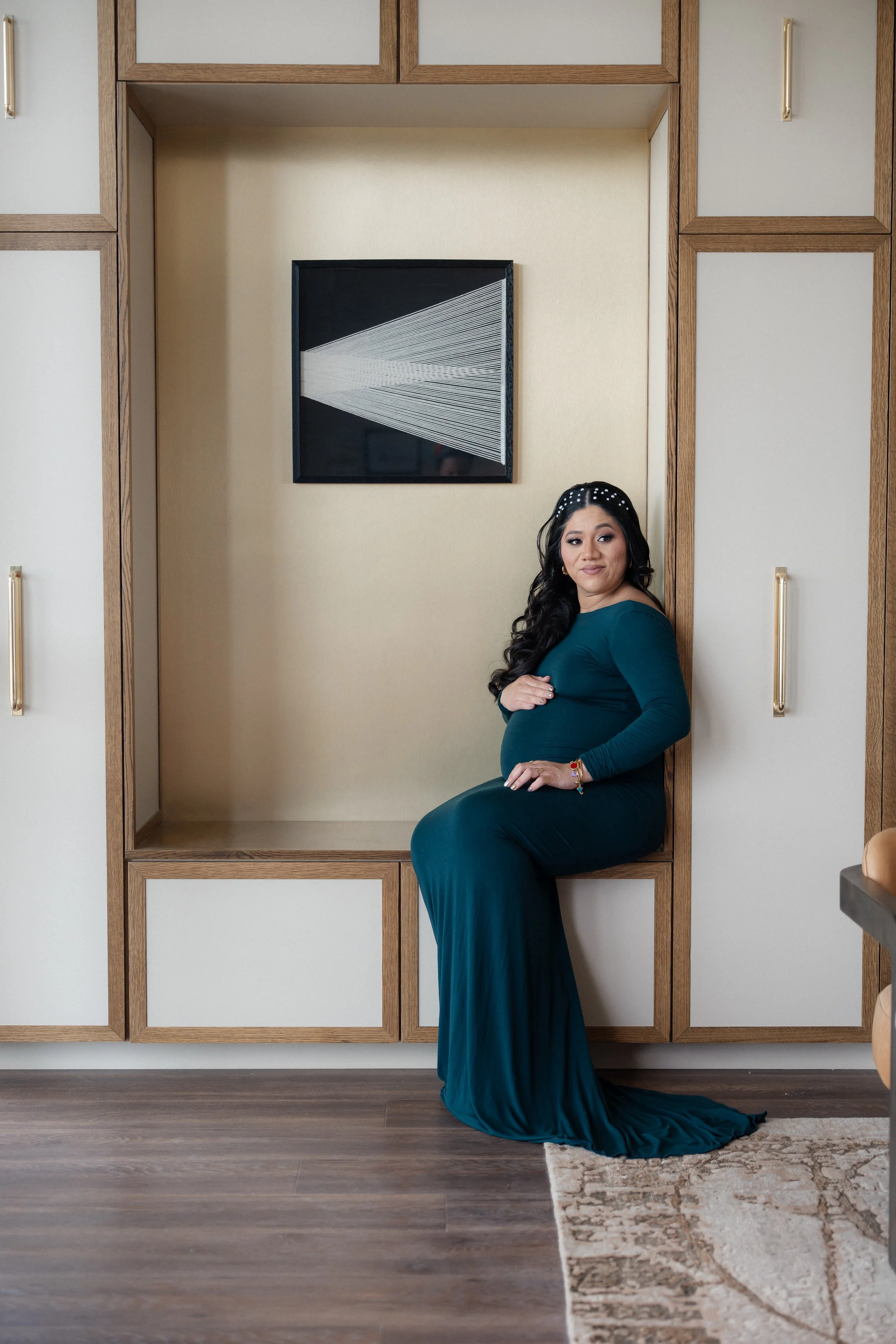 A pregnant woman with long black hair sitting on a ledge in a room with wood-paneled walls and a framed geometric artwork. She is wearing a teal dress, has her arms crossed over her belly, and is smiling slightly. There is a small rug on the wooden f