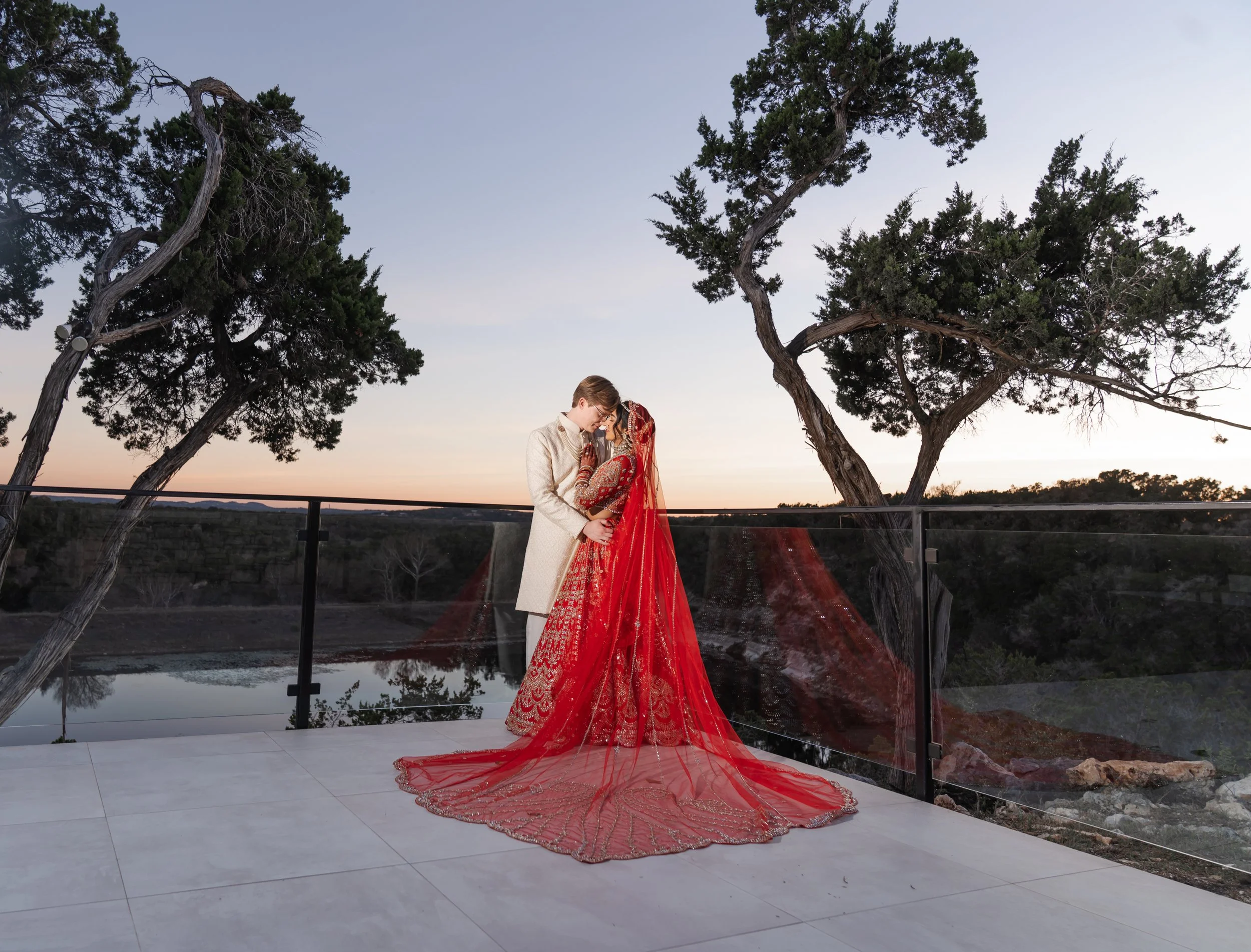 A dramatic photo of couple dressed in traditional Indian wedding attire embracing outdoors during sunset, with tall trees and a reflective water body in the background on the terrace at the Videre Estate.