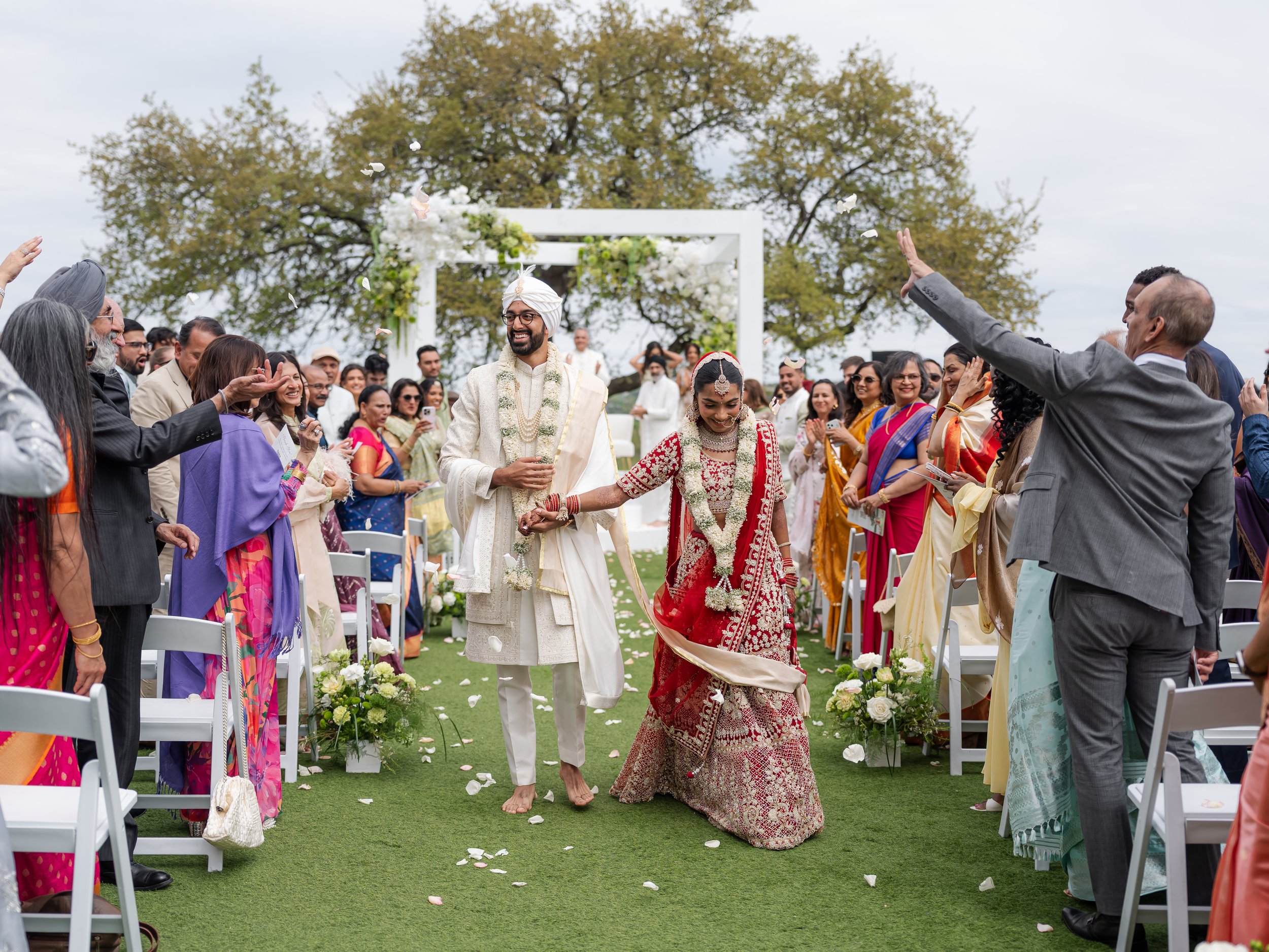 Couple in Indian wedding attire walking down an outdoor aisle with family.