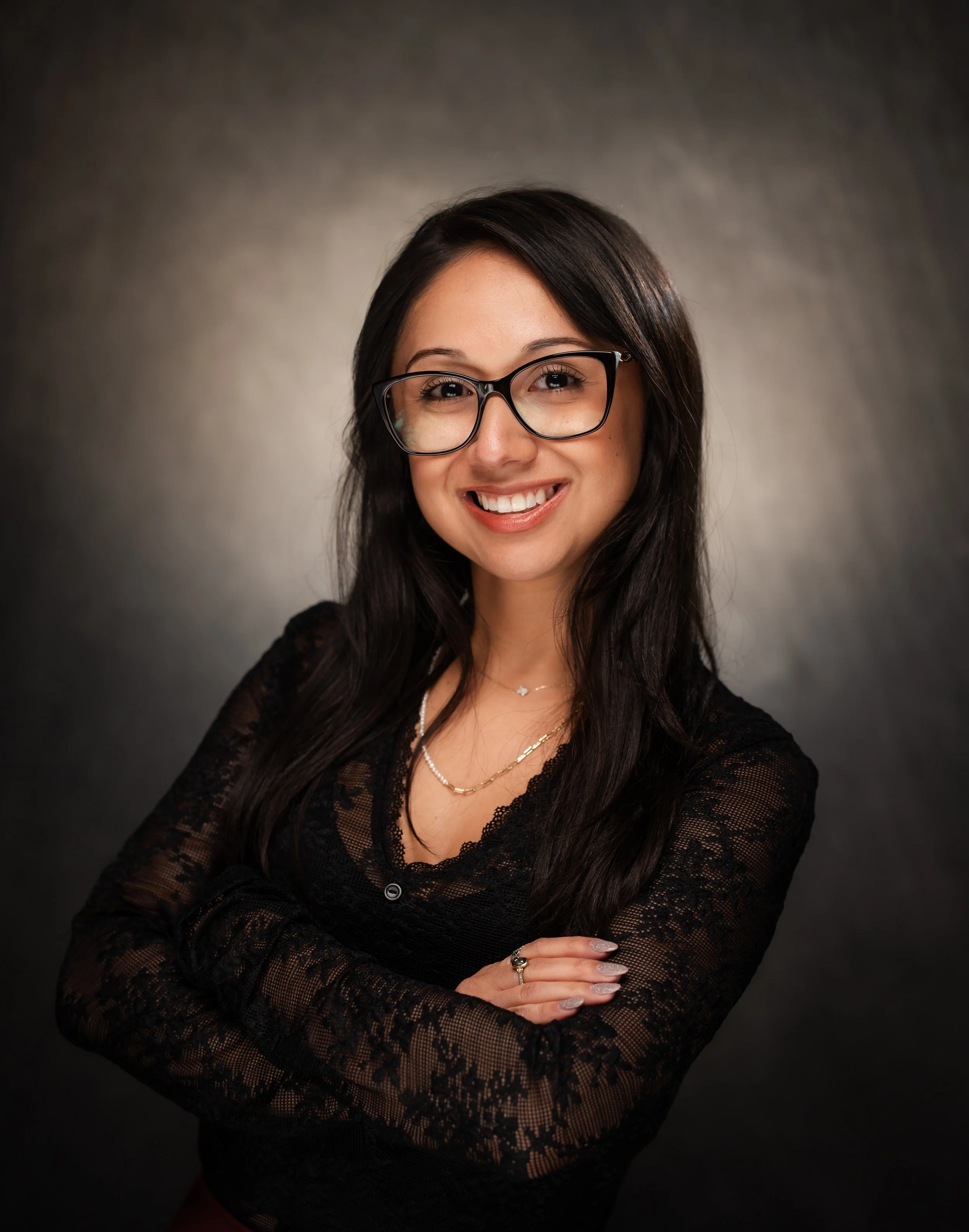 Smiling woman with glasses in a black lace top, posed against a dark background.