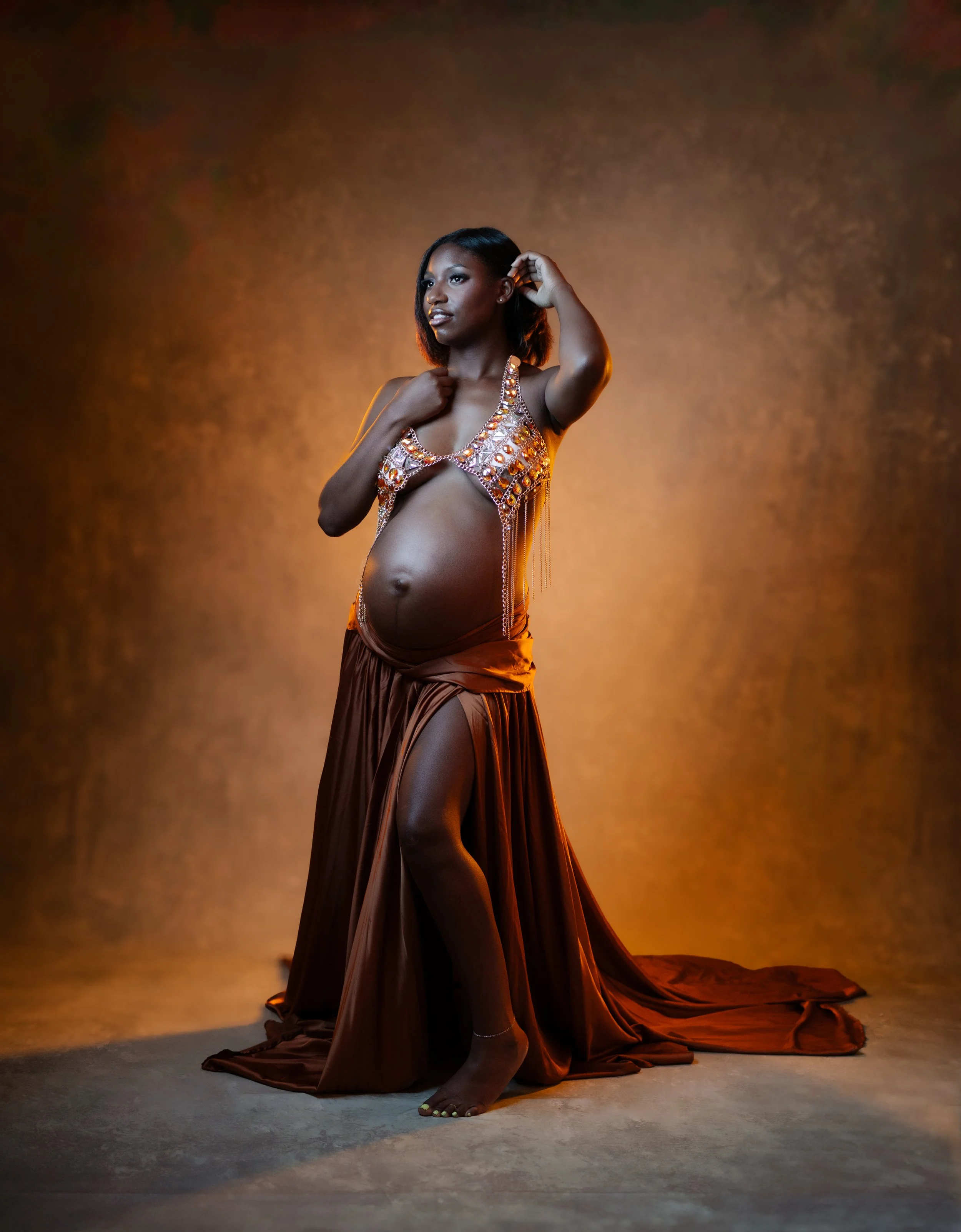 A woman standing in a dimly lit studio with a warm brown background. She is wearing a revealing, metallic, bejeweled top with dangling chains and a long, flowing brown skirt. She has black shoulder-length hair and is barefoot, with her left hand touc