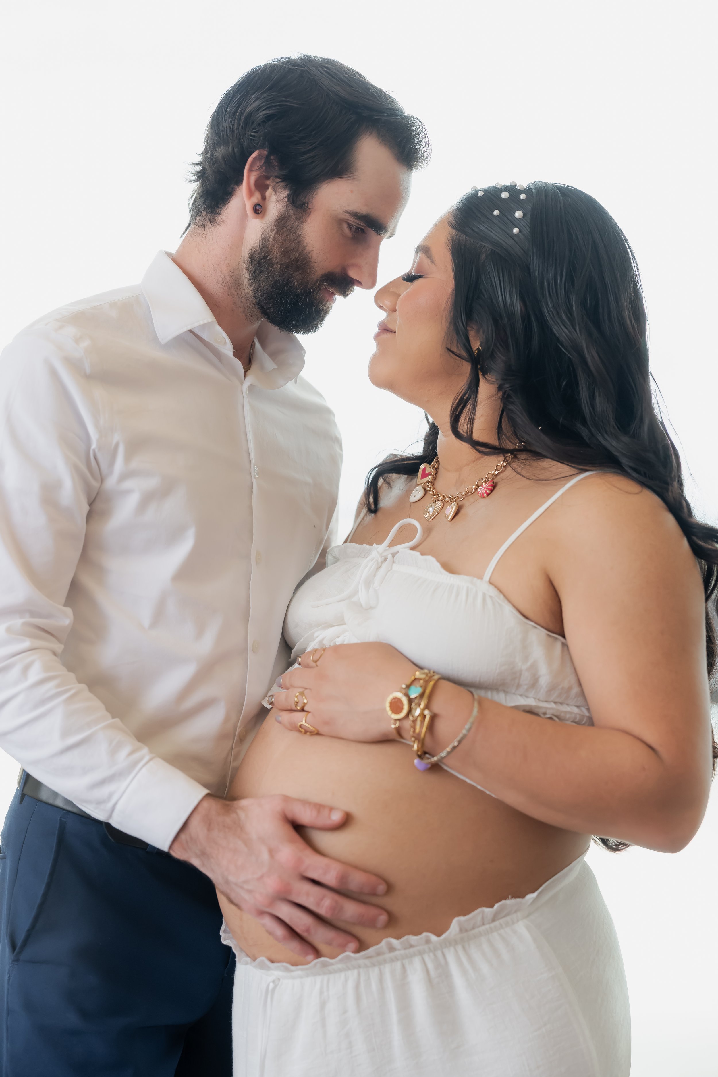 A man with dark hair and a beard gently touching the pregnant belly of a woman with long dark hair, wearing jewelry, both with their eyes closed, in an intimate moment.